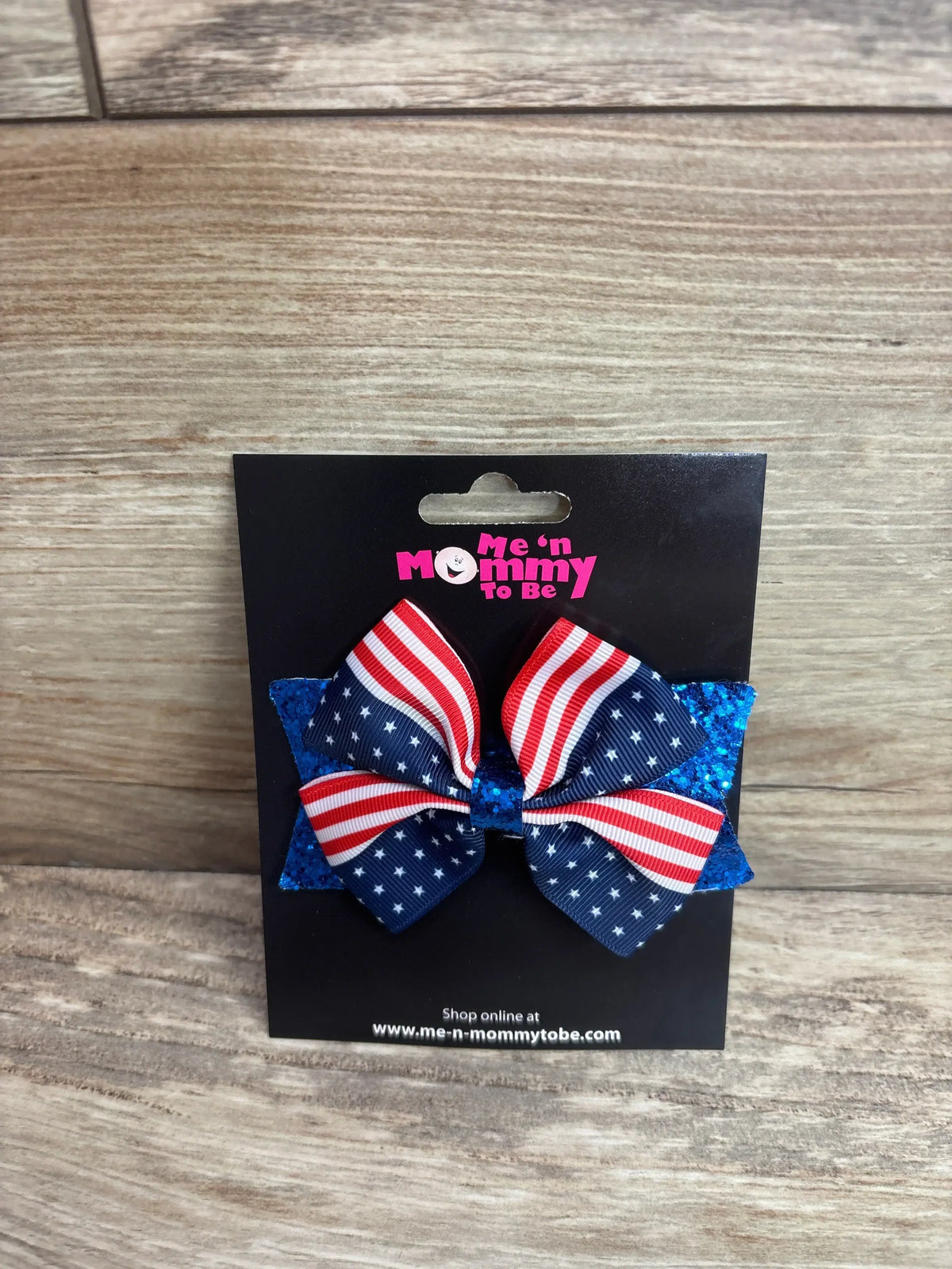 Front view of Fourth of July hair bow showing flag ribbon and blue glitter backing on black card with wood background