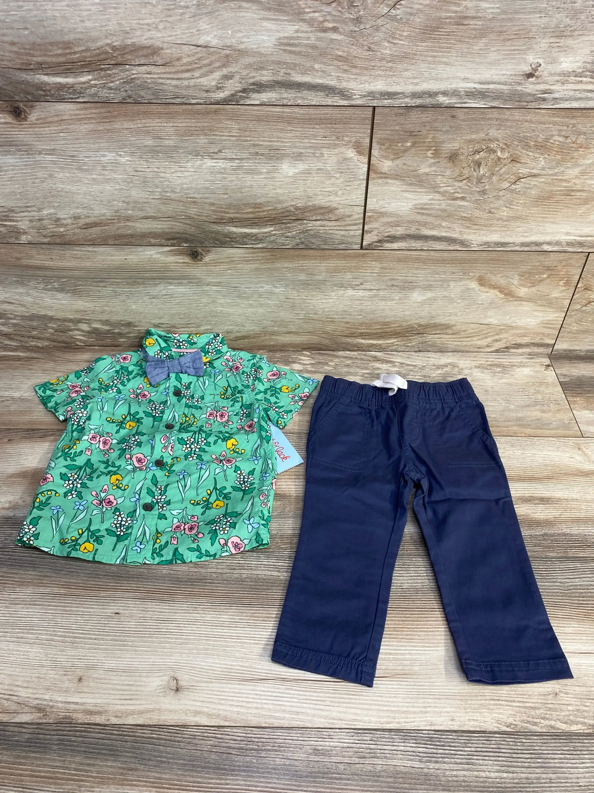 Flat lay of Cat & Jack toddler 3-piece outfit: green floral short-sleeve button-up with blue bow and navy elastic-waist pants on wood floor