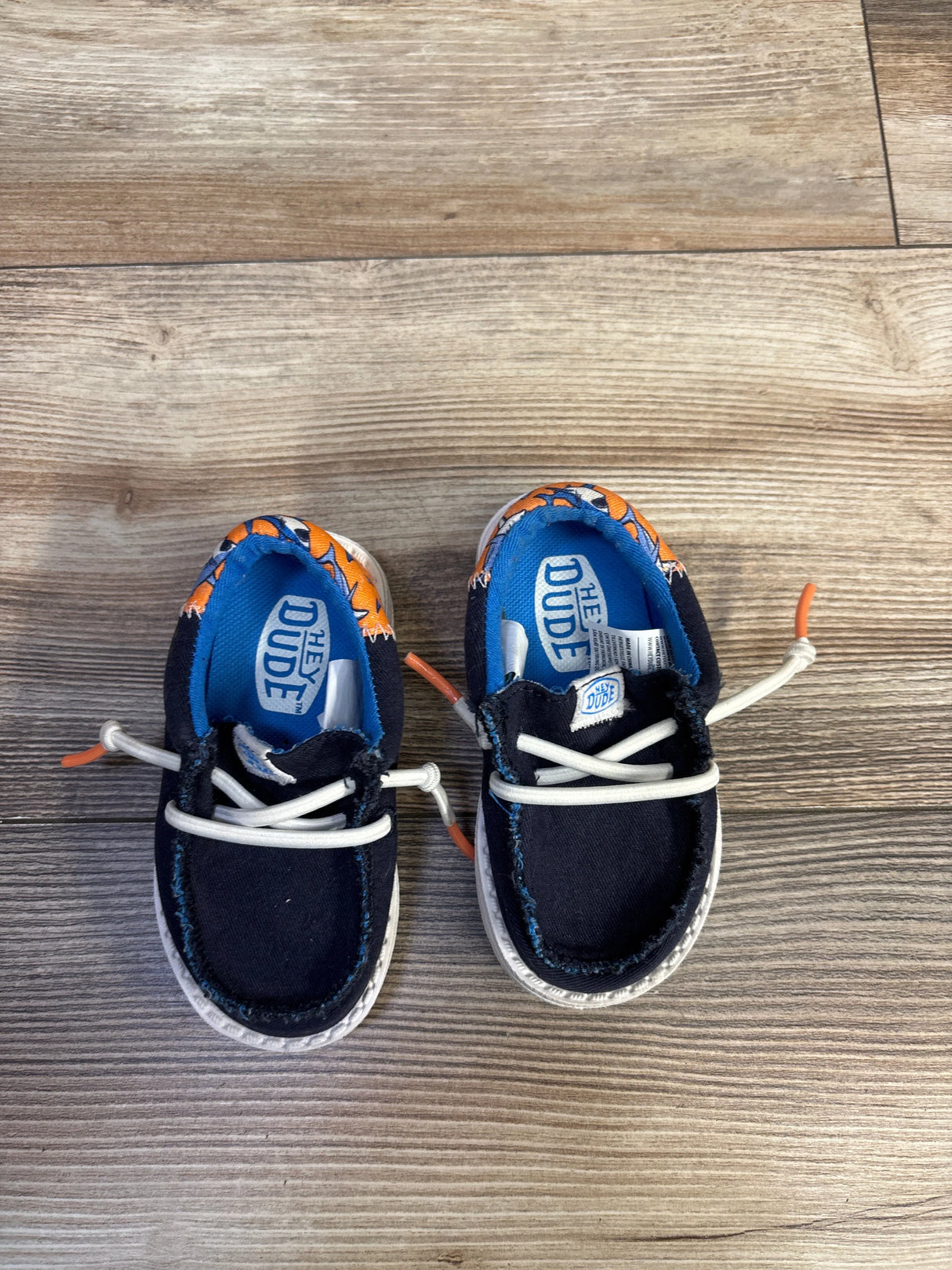 Top-down view of Hey Dude Wally navy kids shoes with white elastic laces and orange shark-print lining on wood floor