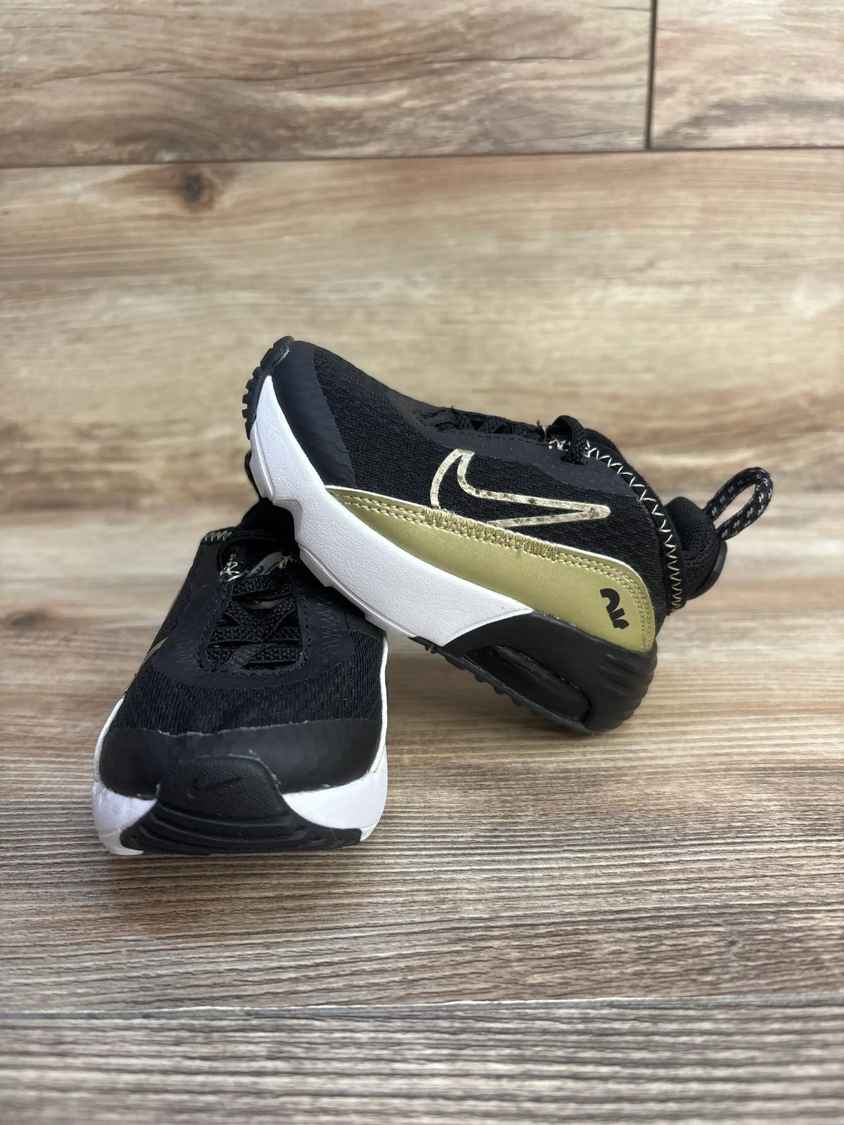 Front three-quarter view of Air Max 2090 TD sneakers size 4C, black knit with metallic gold swoosh and white midsole on wooden floor