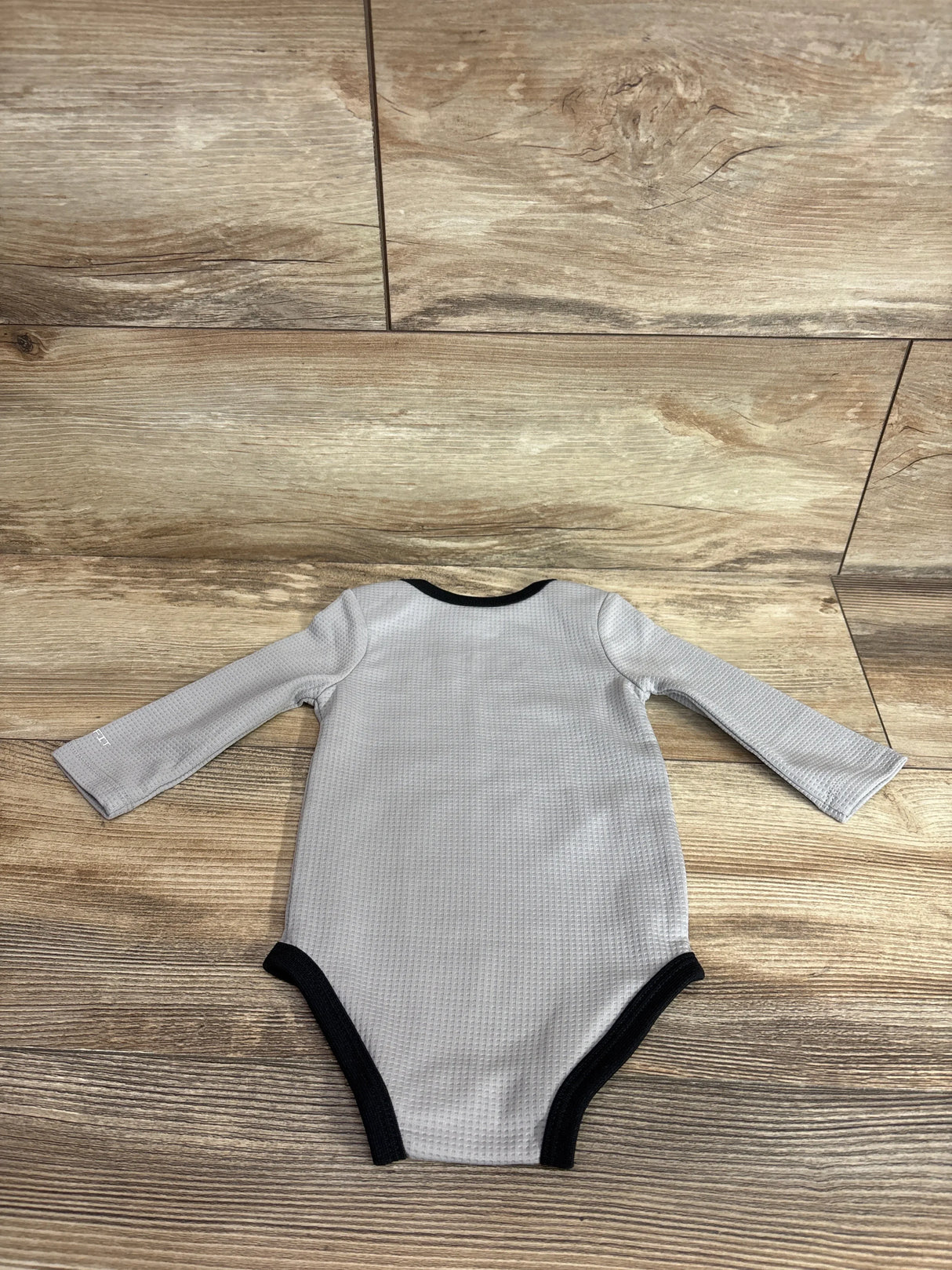 Flat-lay back view of Nike Dri-FIT long-sleeve baby bodysuit showcasing grey waffle texture and black trim on wood floor background