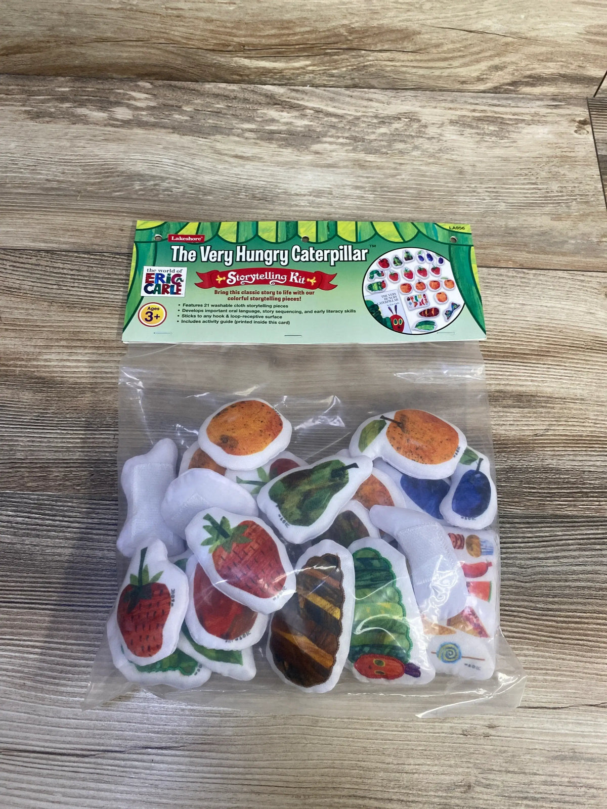 Front view of The Very Hungry Caterpillar storytelling kit with header card and fabric food pieces in clear bag on wood