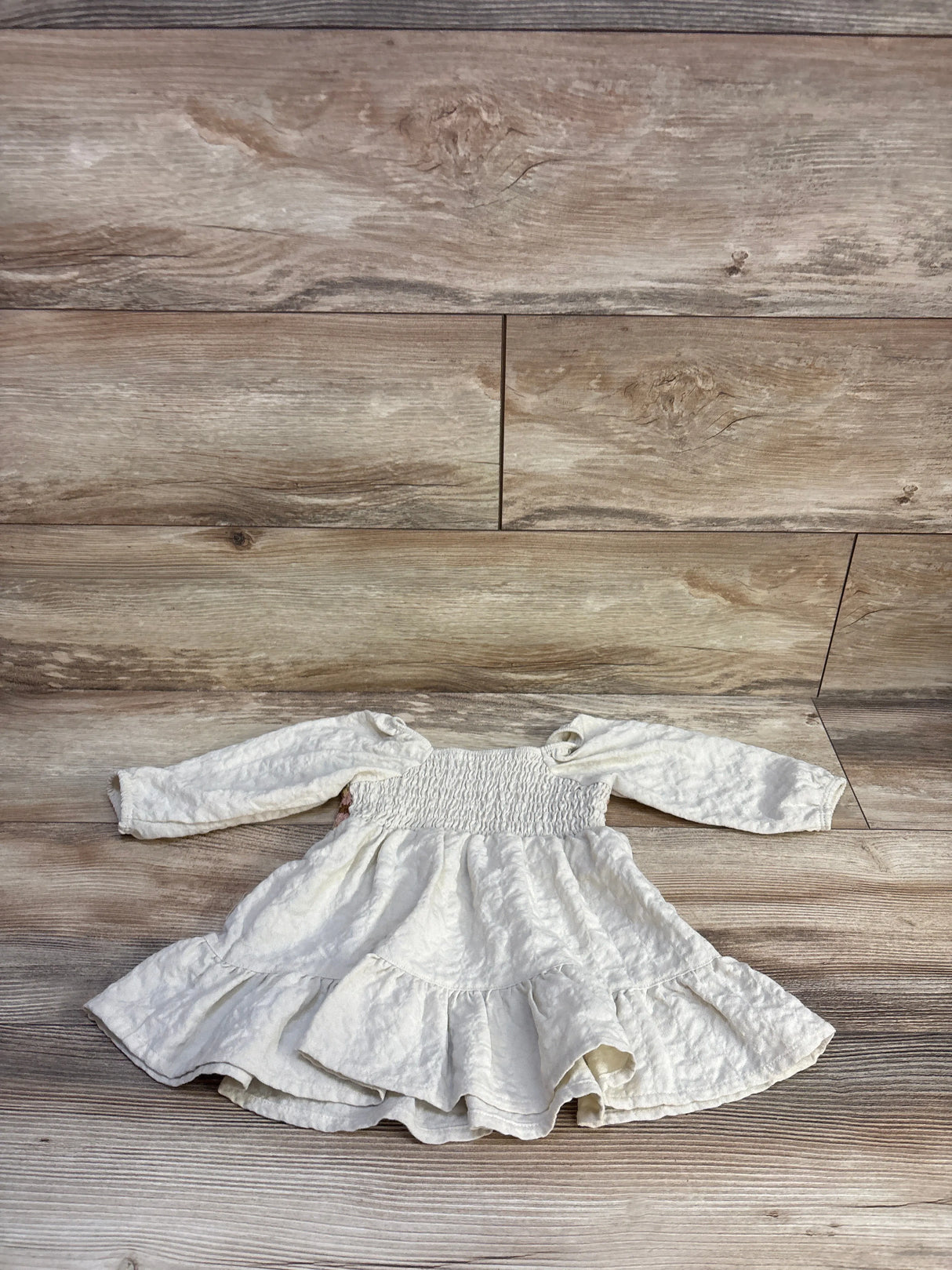 Front flat-lay of beige Little Co crochet baby dress with smocked bodice and tiered ruffle skirt on wood background