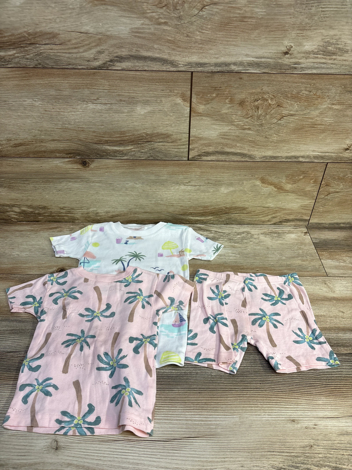 Flat-lay of Just One You 3pc pajama set 3T with pink palm print top, shorts, and white beach-print tee on wood floor