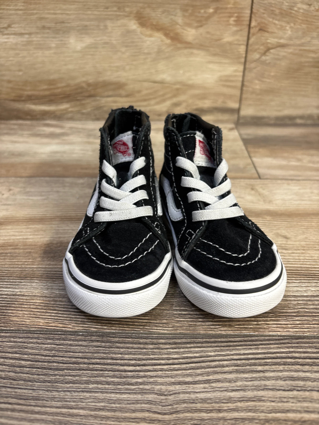 Front view of toddler black Vans Sk8-Hi Zip sneakers showing white laces, white side stripe, and high-top silhouette on a wood floor