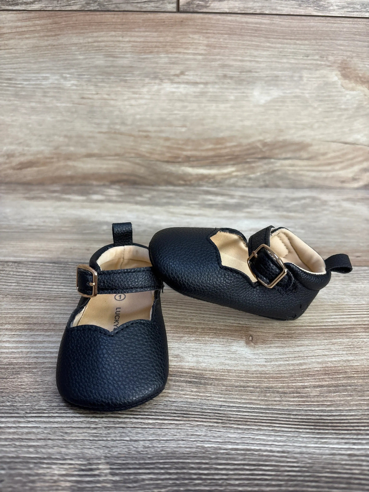 Lucky Brand black leather Mary Jane crib shoes with gold buckles on a wood surface