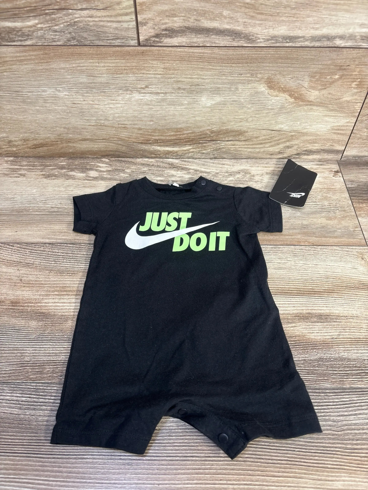 Flat lay of Nike shortie romper showcasing black cotton fabric and neon green “JUST DO IT” logo on wood floor background