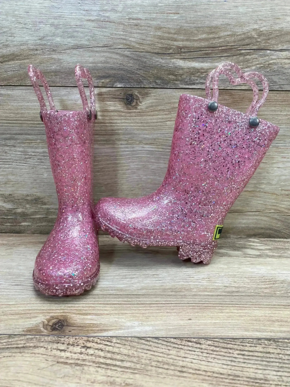 Side and front view of toddler pink glitter Western Chief rain boots with heart shaped pull handles on wood background