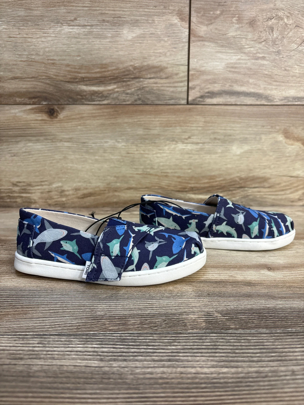 Kids' shark print slip-on shoes, navy blue with colorful fish, white soles, on wood floor