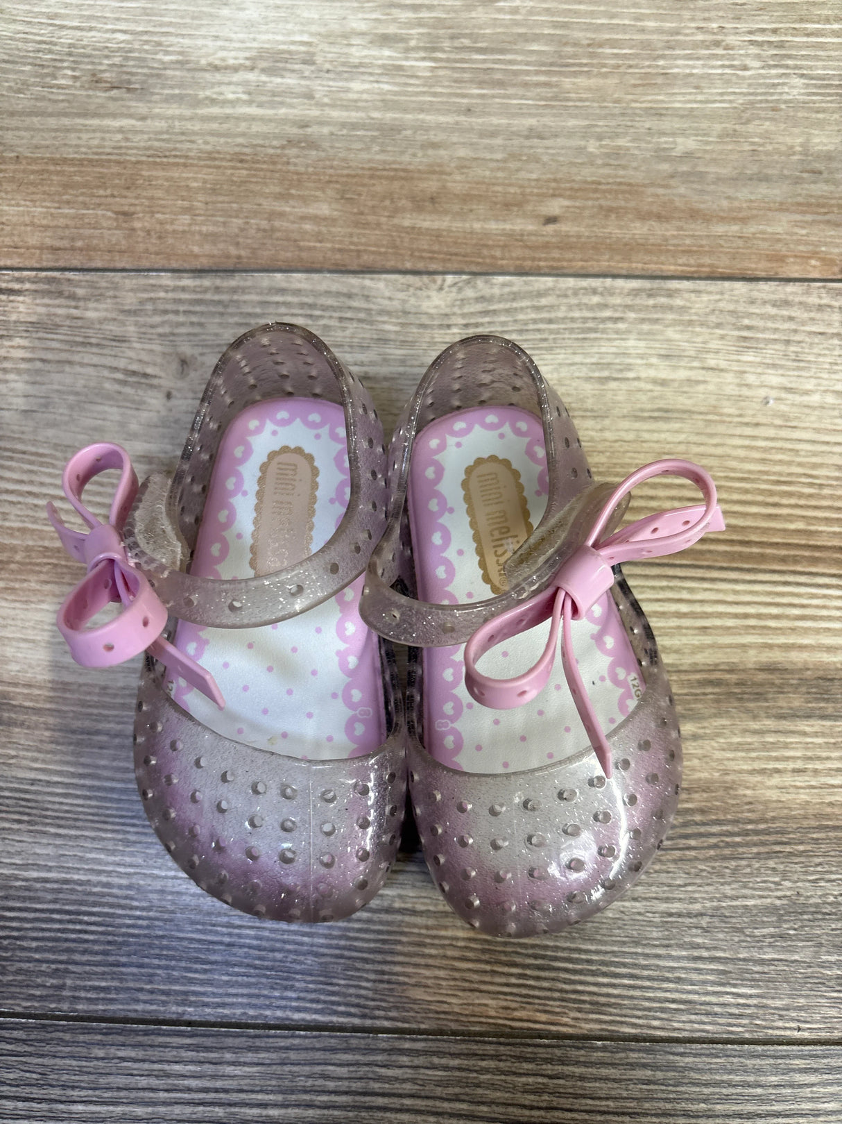 Top view of Mini Melissa Ultragirl jelly Mary Jane shoes showing silver glitter finish, pink bow straps, and scalloped pink insoles on wooden floor background