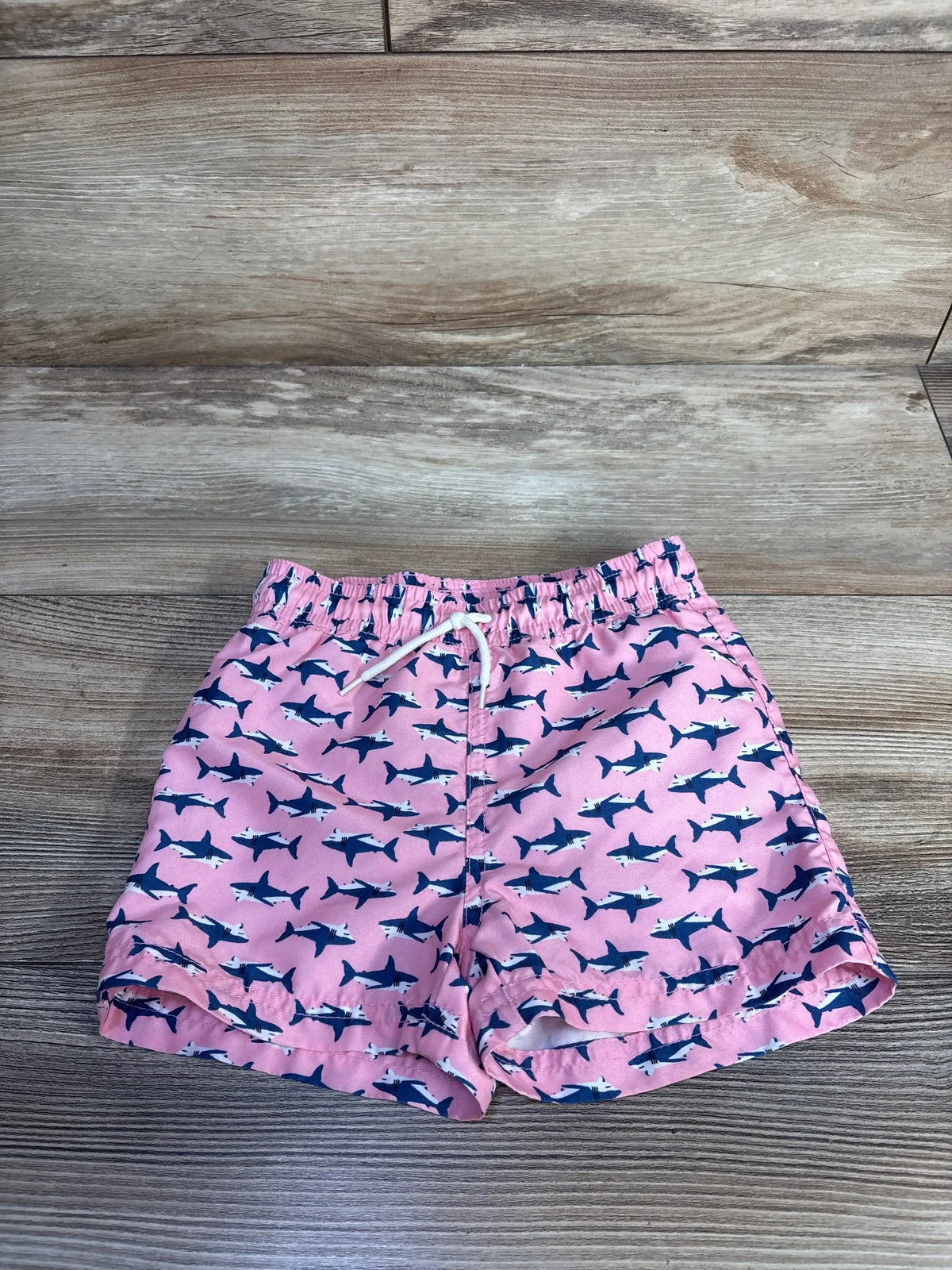 Front view of pink Janie & Jack toddler swim trunks with navy shark print, elastic waistband and white drawstring on wood background