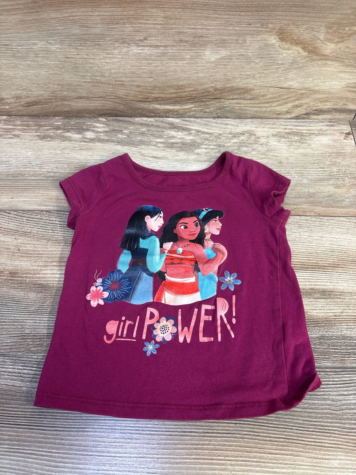 Flat lay of mauve Disney Girl Power toddler shirt with front graphic of three Disney characters and girl power text on wood background