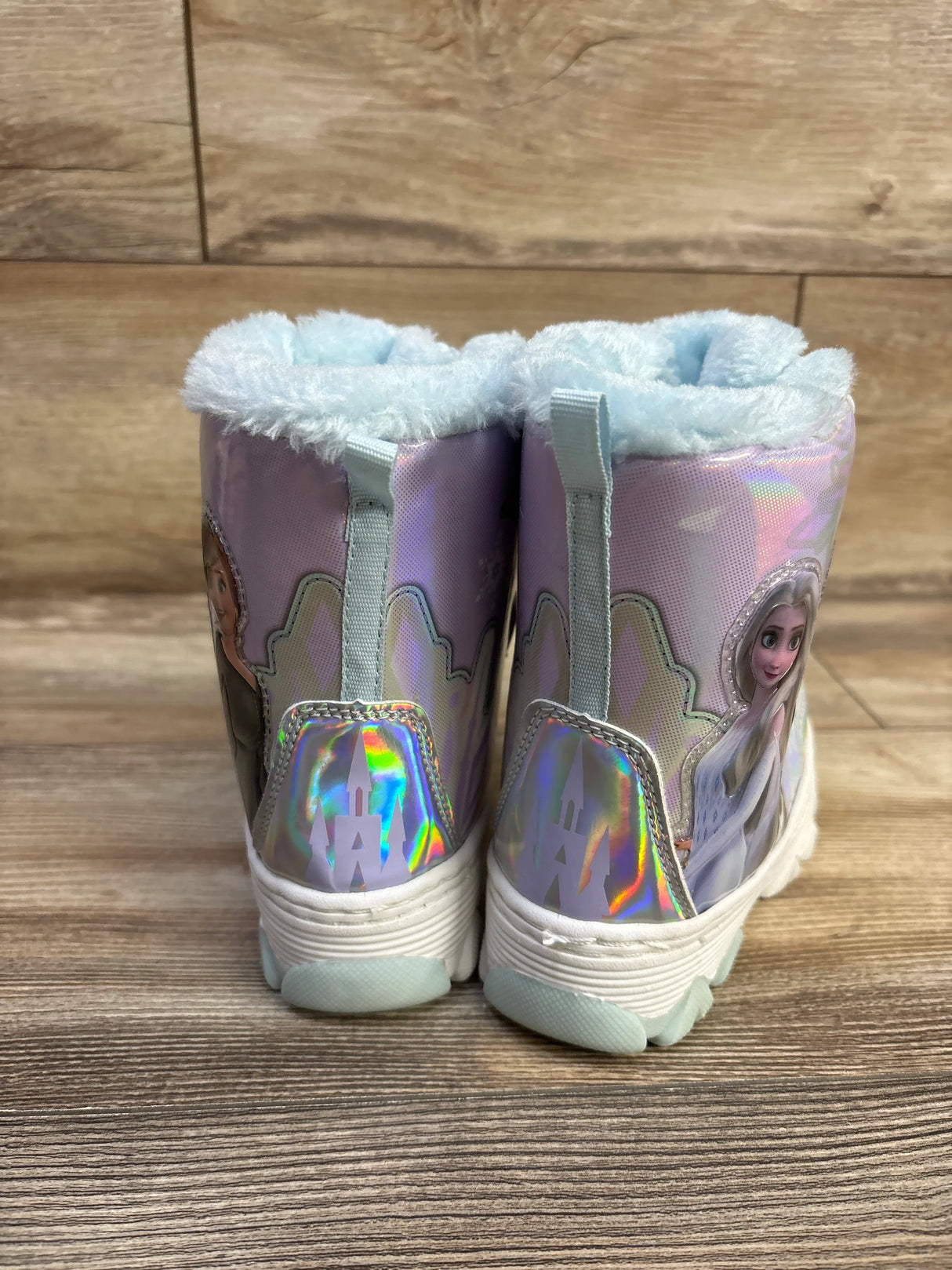 Rear view of Disney Frozen silver iridescent snow boots showing blue faux-fur cuff and holographic castle on wood floor