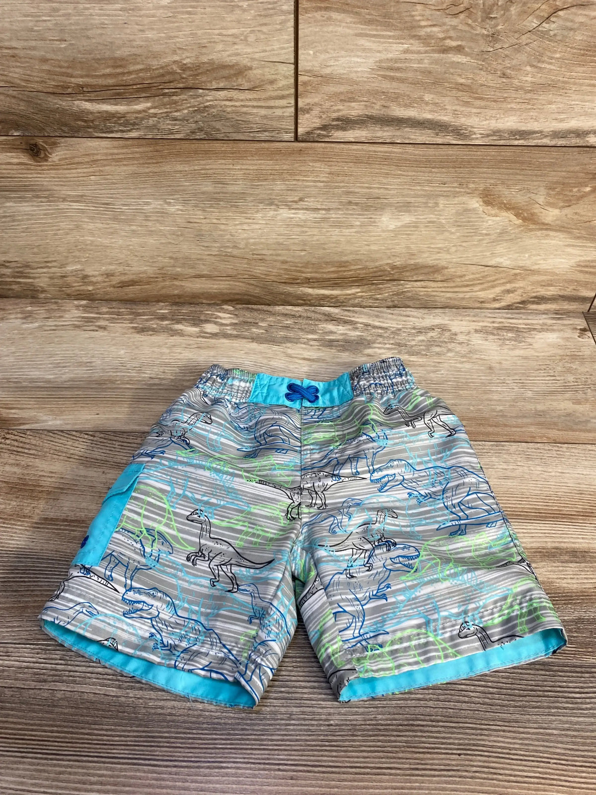 Front view of UV Skinz swim trunks in grey showcasing dinosaur print and turquoise drawstring waistband flat lay on wood plank floor