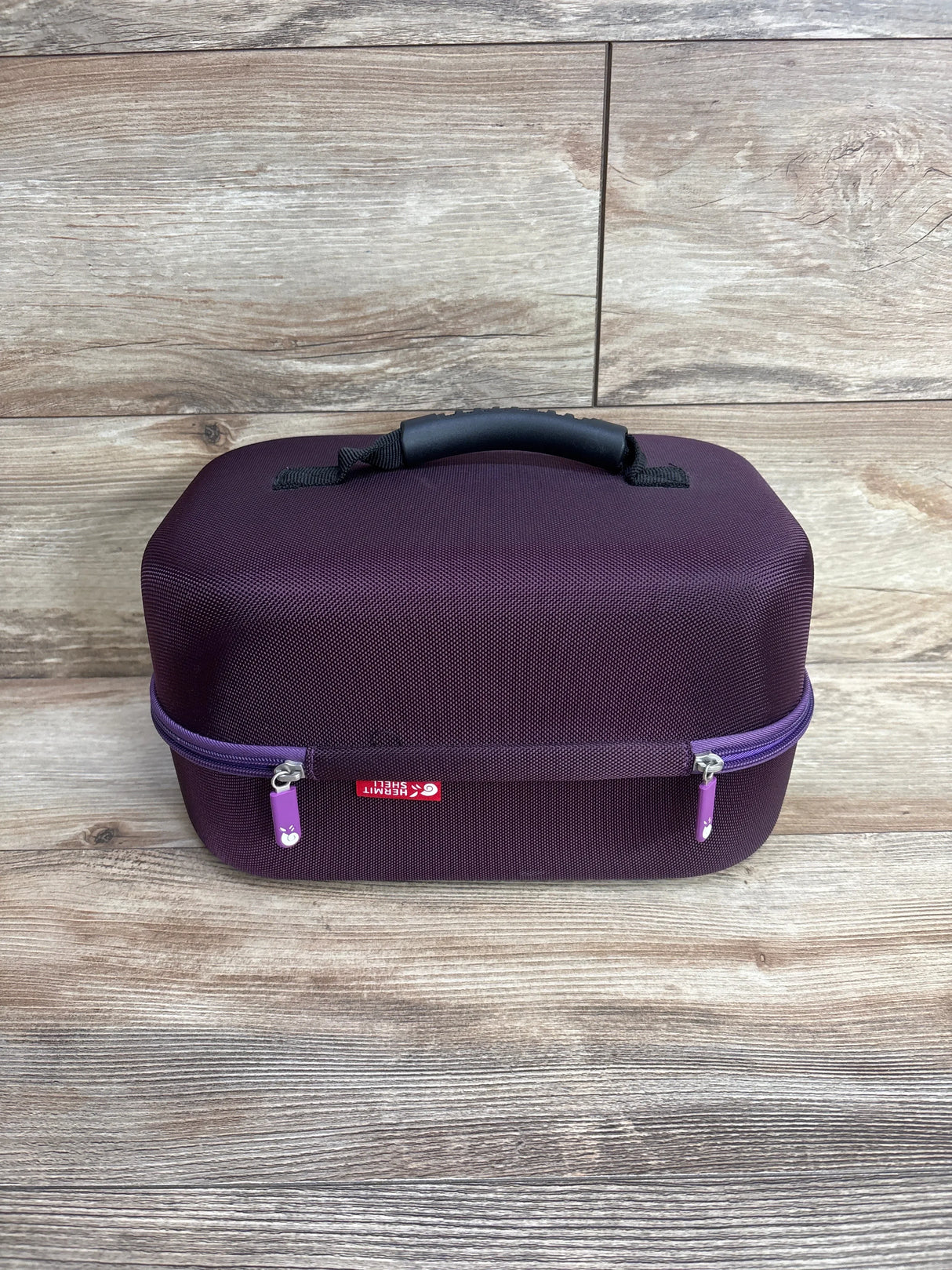 Front view of Hermitshell hard EVA travel case for Spectra breast pump with textured purple shell, black padded handle and purple zipper on wood background
