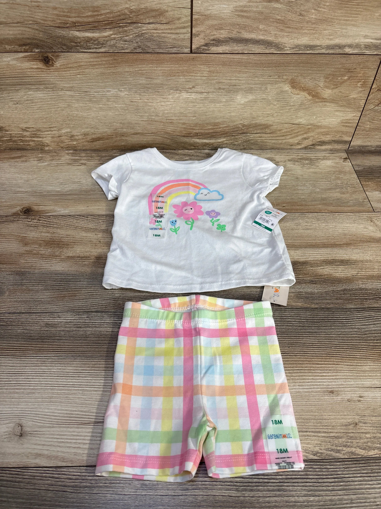 Flat lay of Garanimals 2-piece baby set size 18M: white tee with rainbow flower graphic and plaid shorts on wood background