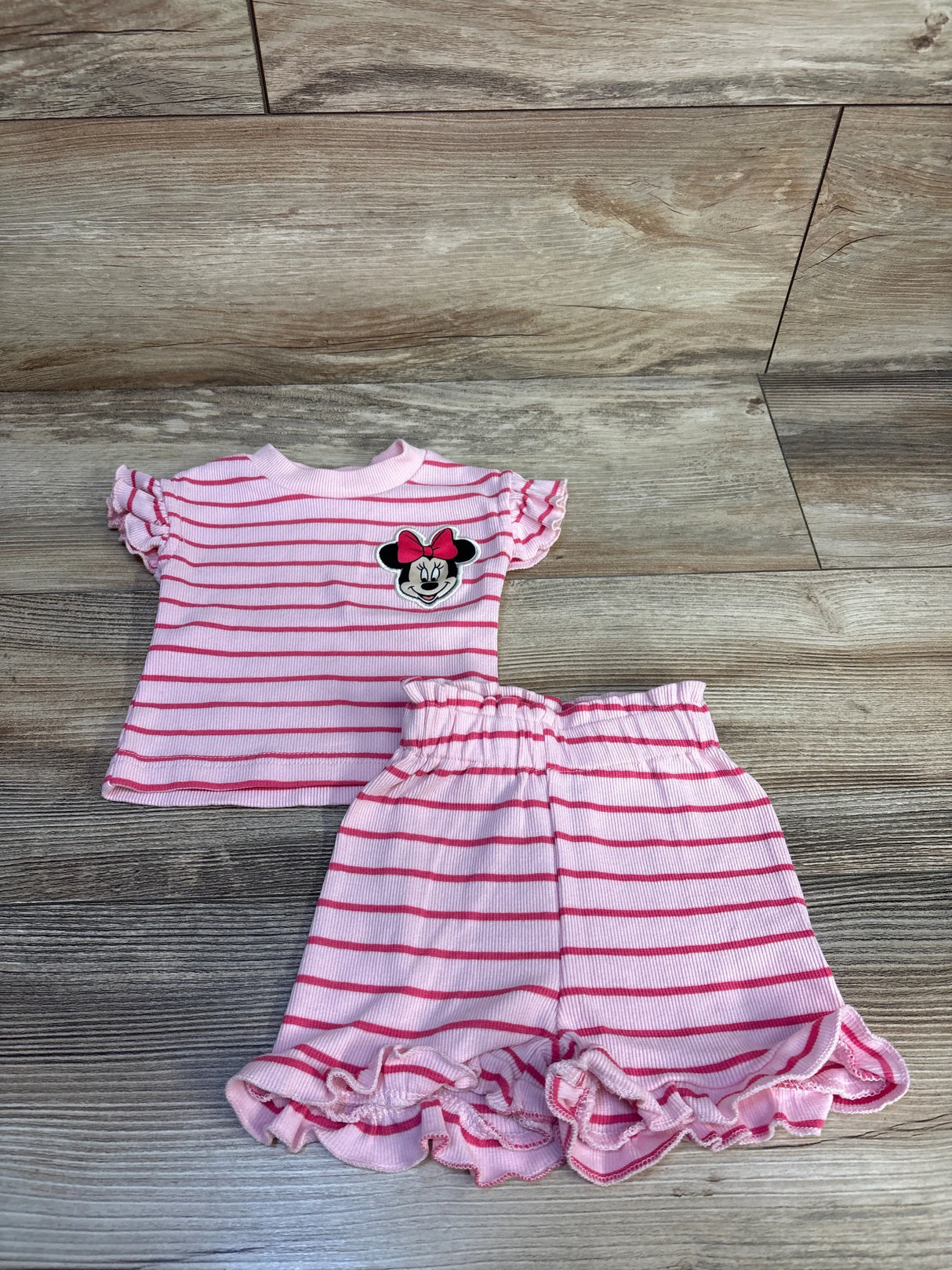 Flat lay of Disney Baby 2pc set showcasing pink striped Minnie Mouse top with ruffle sleeves and ruffle-hem shorts on wood plank floor