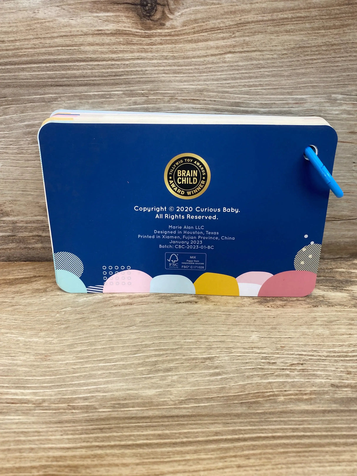 Back view of Curious Baby Activity Cards showing navy cover with Brain Child award seal, blue ring, and colorful edge on wood