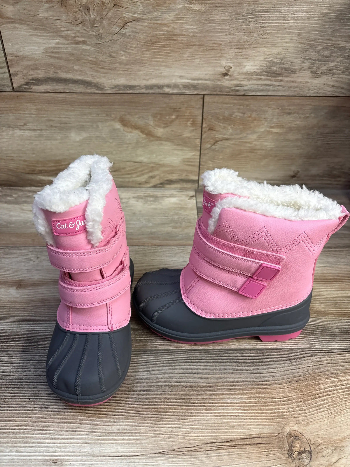 Front three-quarter view of Cat and Jack pink toddler winter boots size 12C showcasing white faux-fur lining, double Velcro straps, and gray rubber toe on wood background
