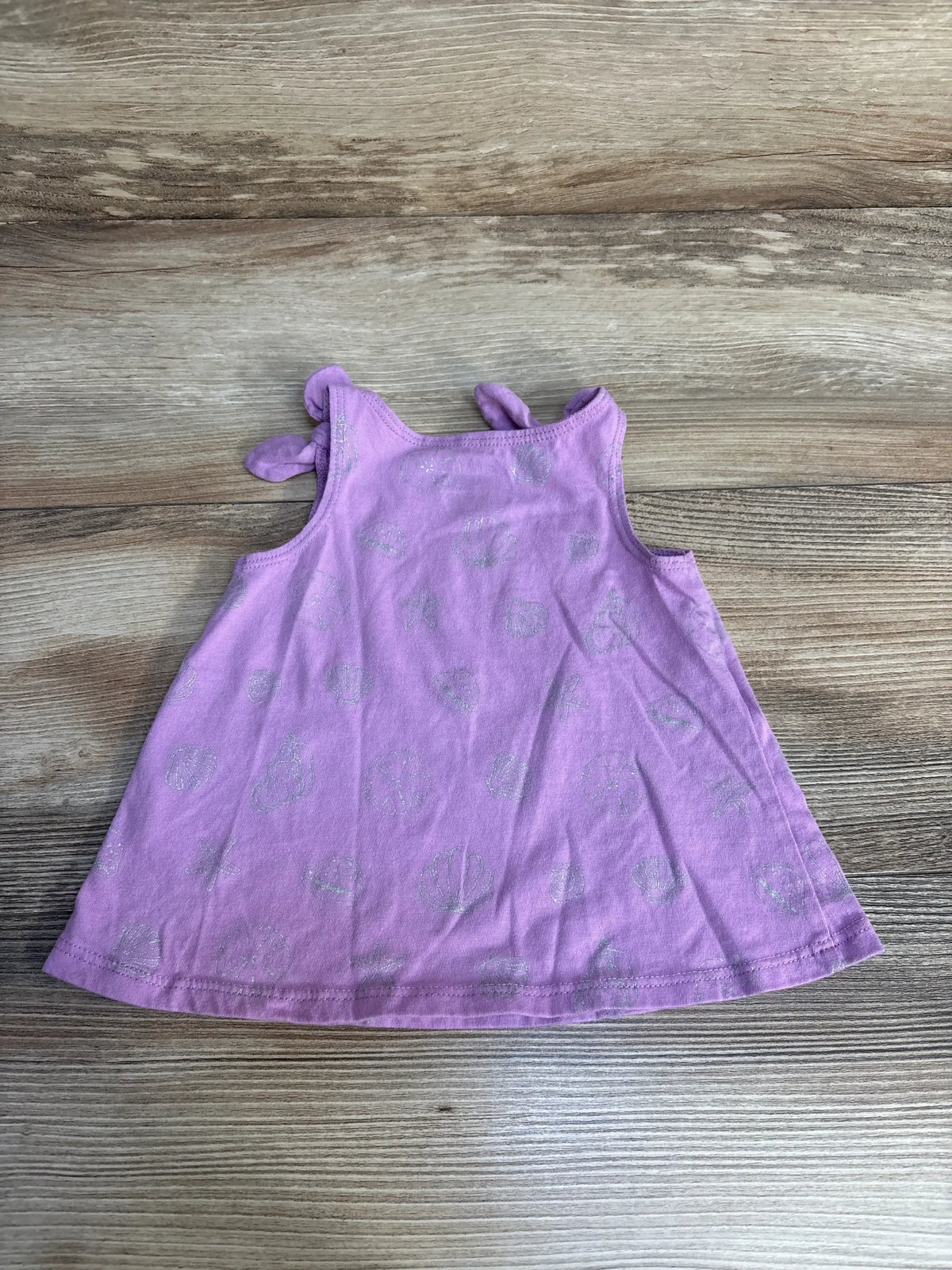 Flat lay of baby purple tank top showcasing knotted shoulder straps and silver glitter seashell print on wood background