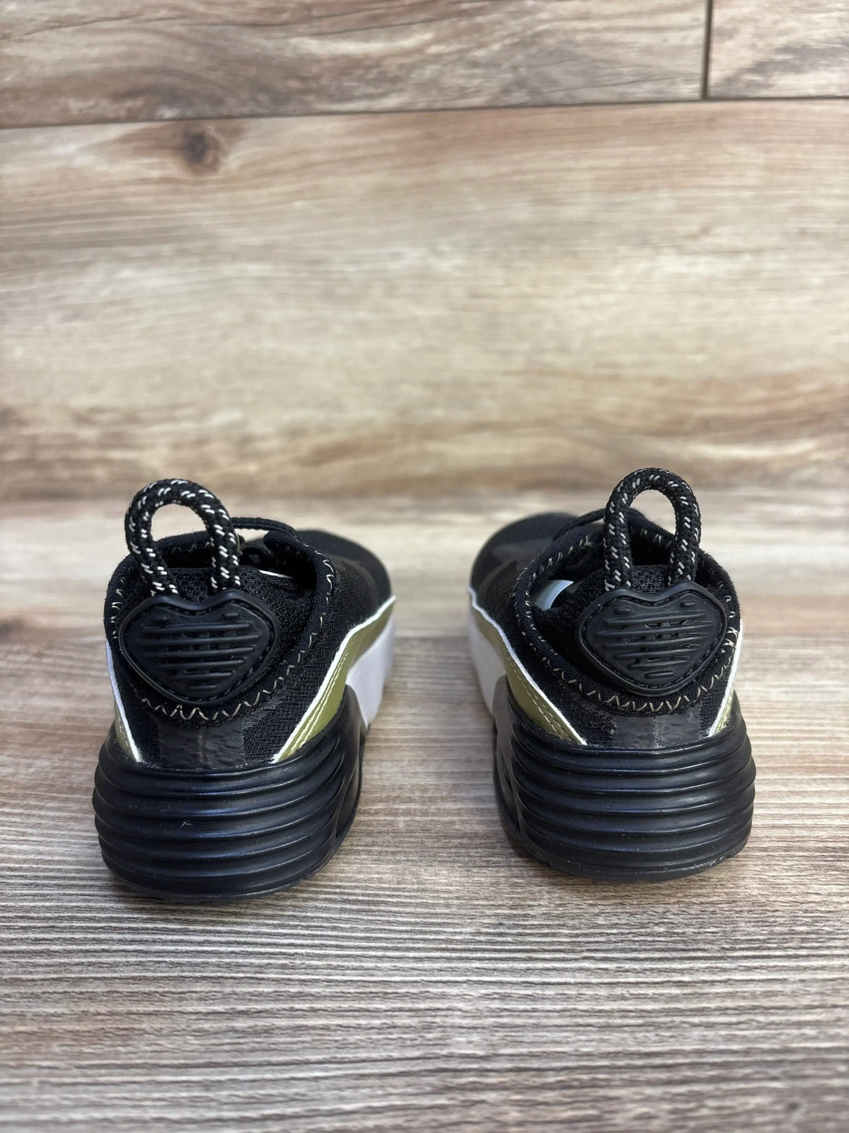 Rear view of Air Max 2090 toddler sneakers in black with metallic gold star, heart-shaped heel loops on wood floor
