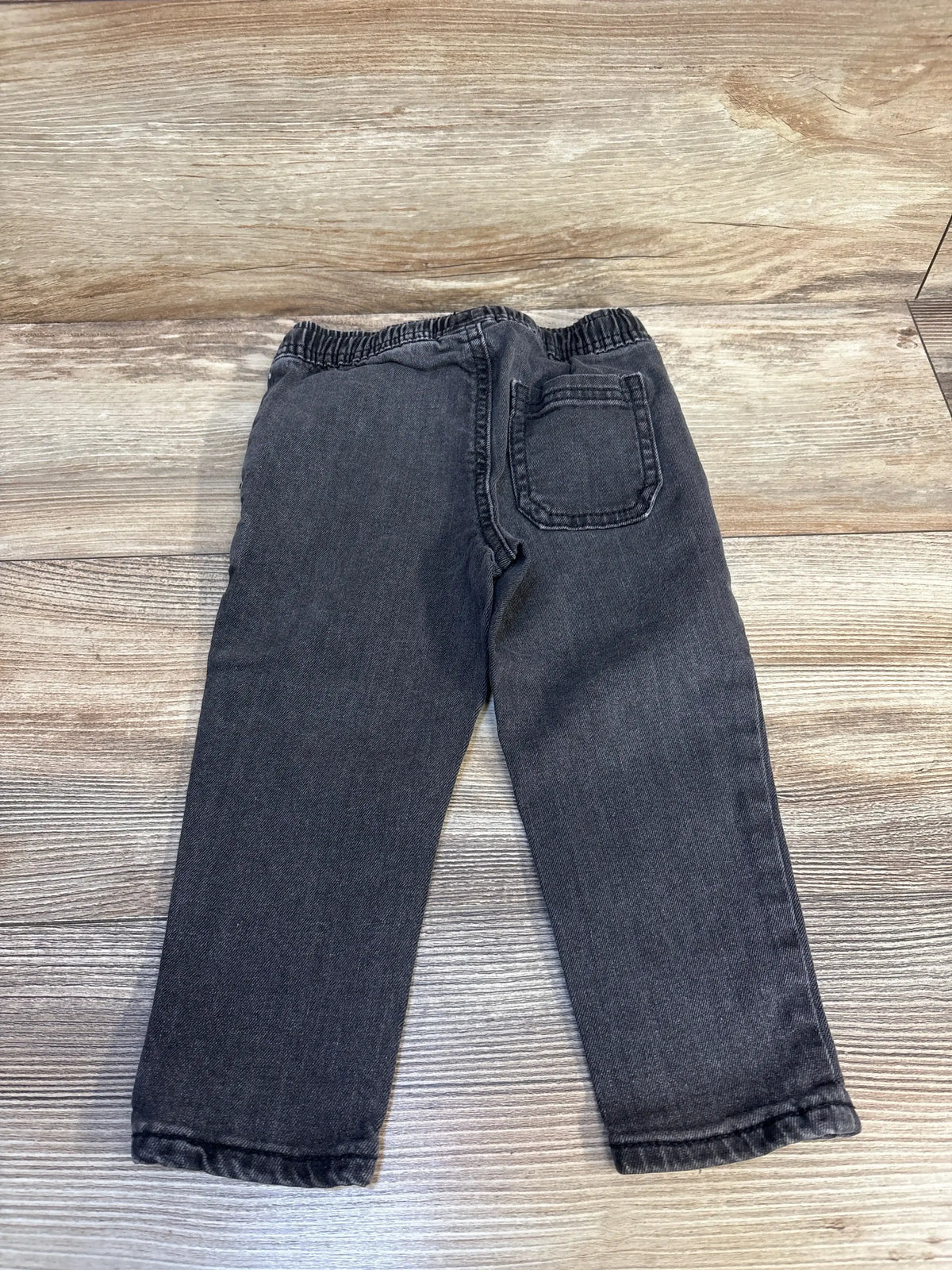 Black denim kids jeans with elastic waistband and back pocket, laid flat on wood floor