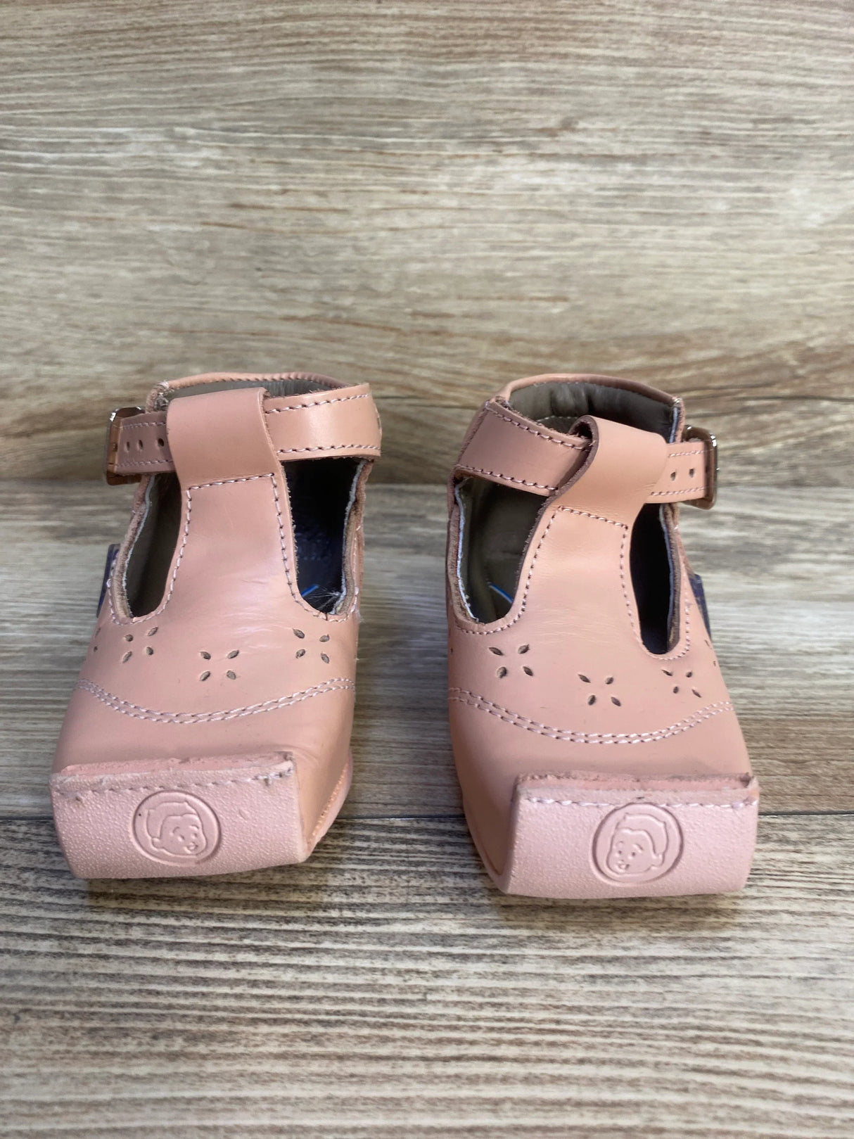 Pink leather toddler shoes with buckle straps and floral cutout design on wood background