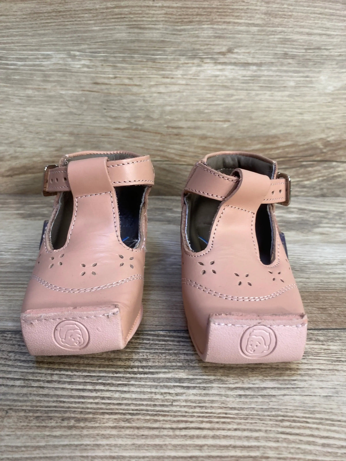 Pink leather toddler shoes with buckle straps and floral cutout design on wood background