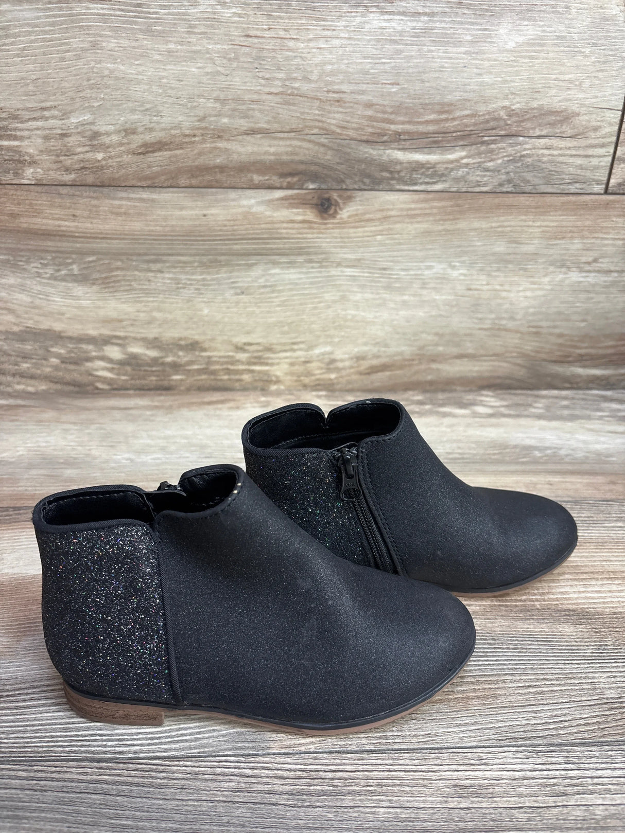 Black toddler ankle boots with side zipper and glitter heel panel on wood background