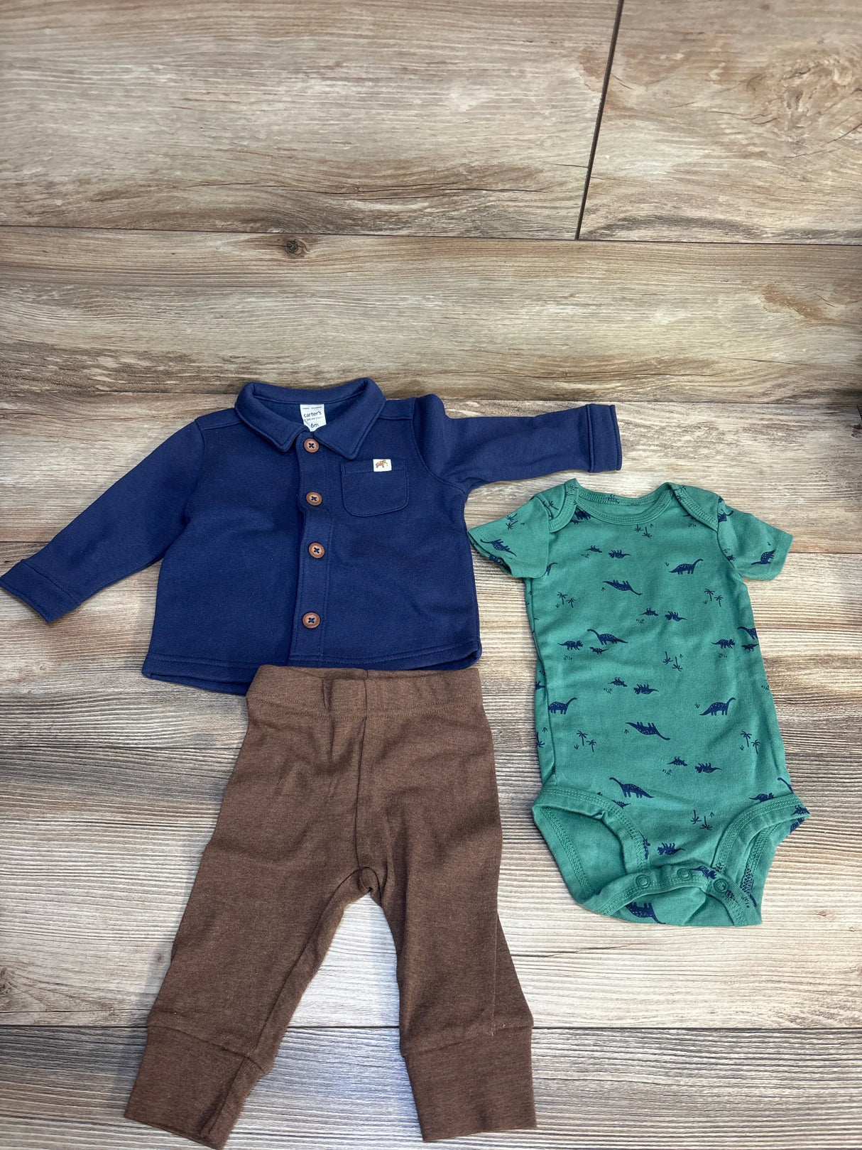 Baby clothes set with blue jacket, brown pants, and green dinosaur print bodysuit on wood floor