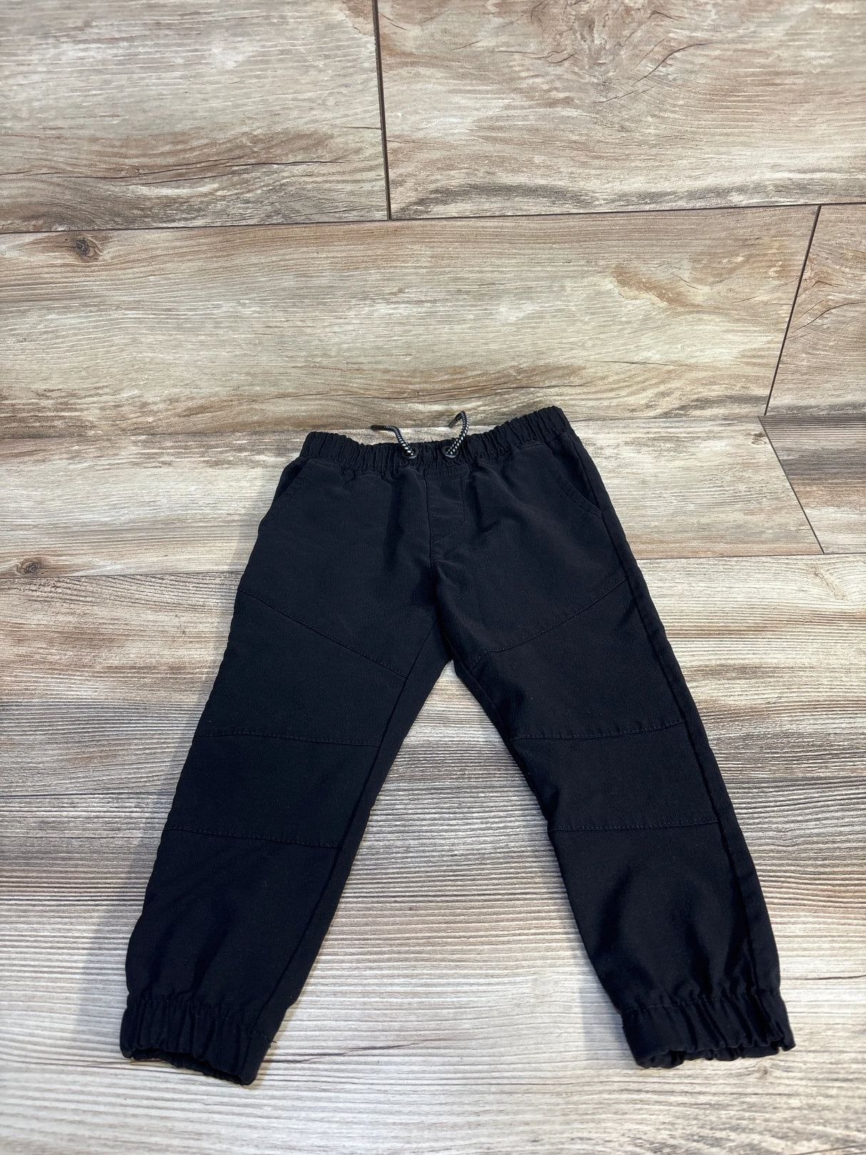 Black kids jogger pants with elastic waist and cuffs, laid on wood floor