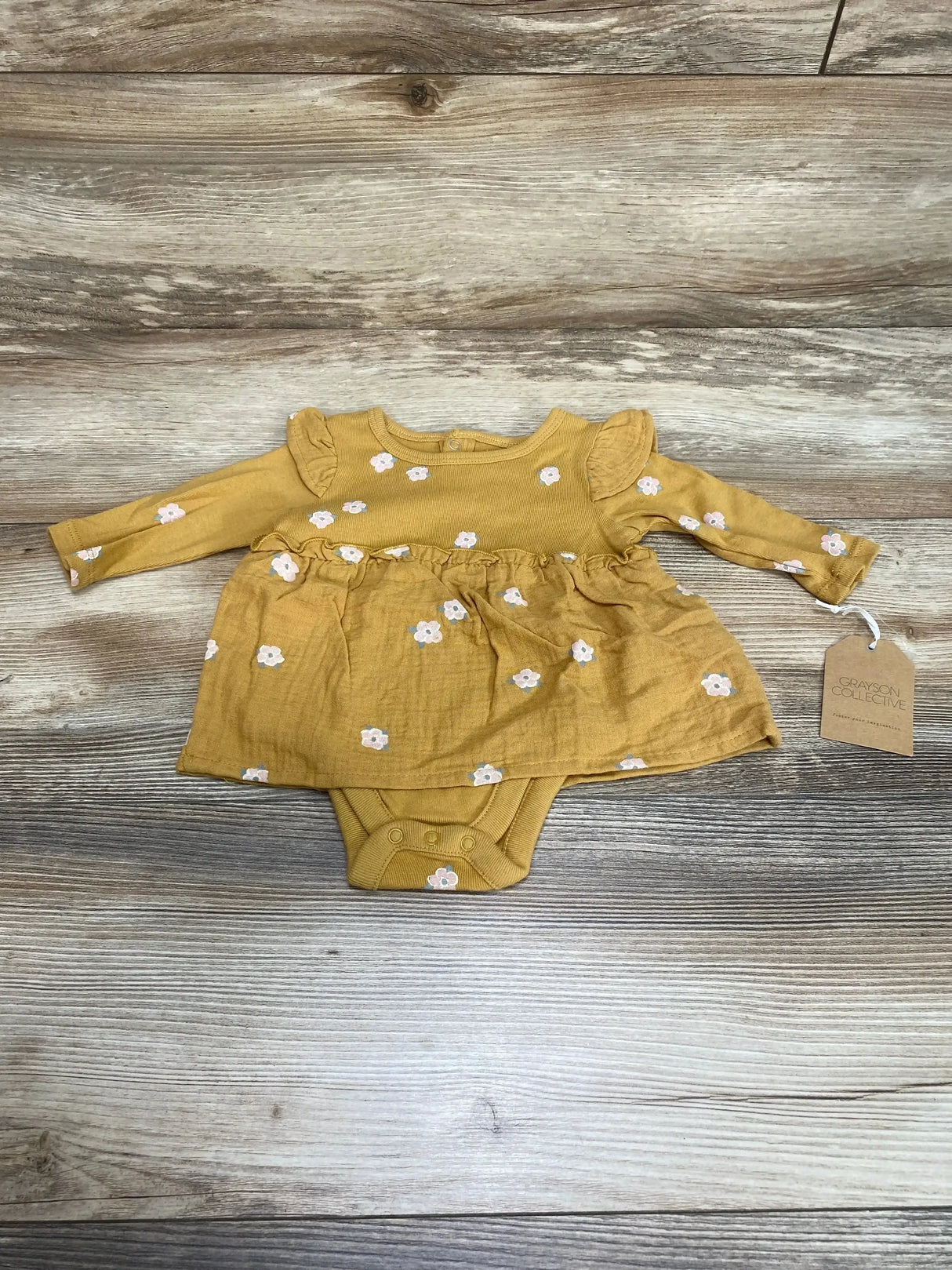 Flat lay of new Grayson Collective Floral Bodysuit Dress in mustard yellow size 0-3m with white daisy print and hangtag on wood plank floor