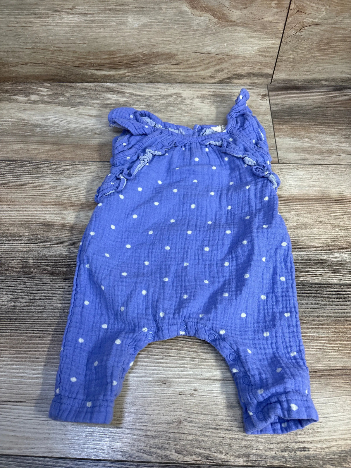 Front flat lay of purple polka-dot gauze baby jumpsuit showing ruffle shoulder straps and snap crotch on wood background