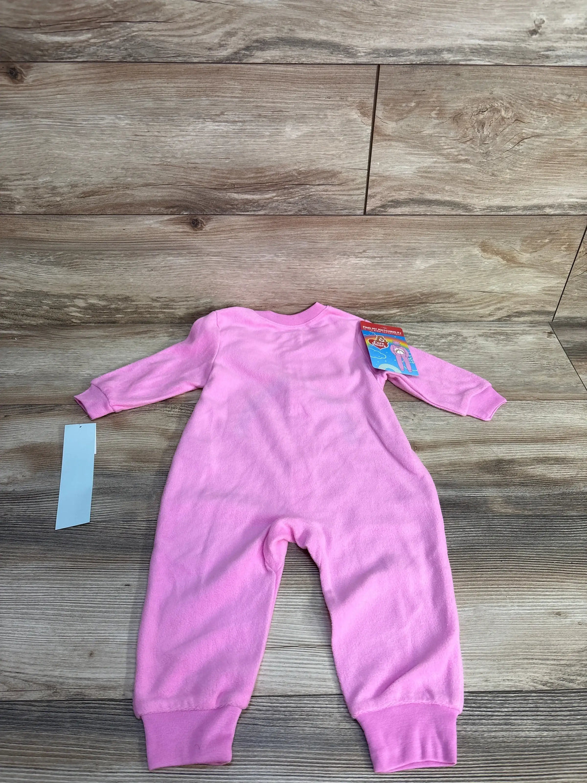 Back view of pink Care Bears sleeper laid flat showing long sleeves, ribbed ankle cuffs, and attached tag on wood background