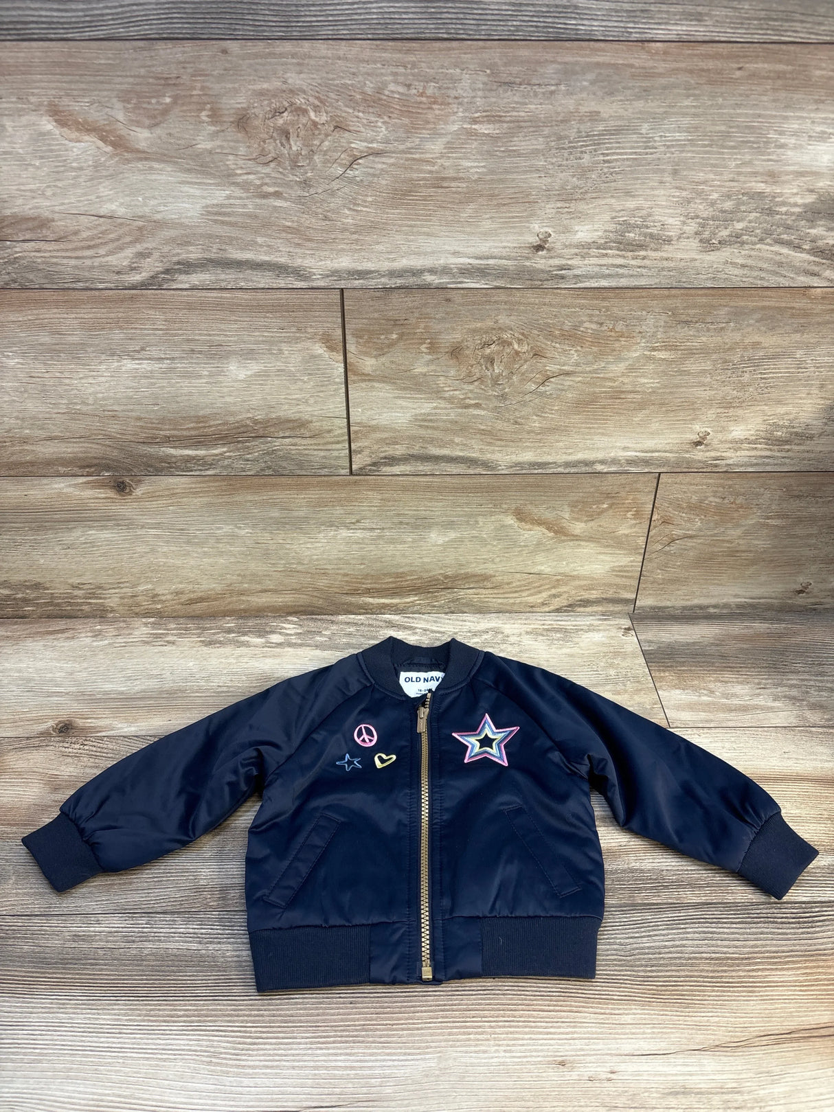 Navy blue toddler bomber jacket with embroidered patches, zip front, and ribbed cuffs on wood floor.