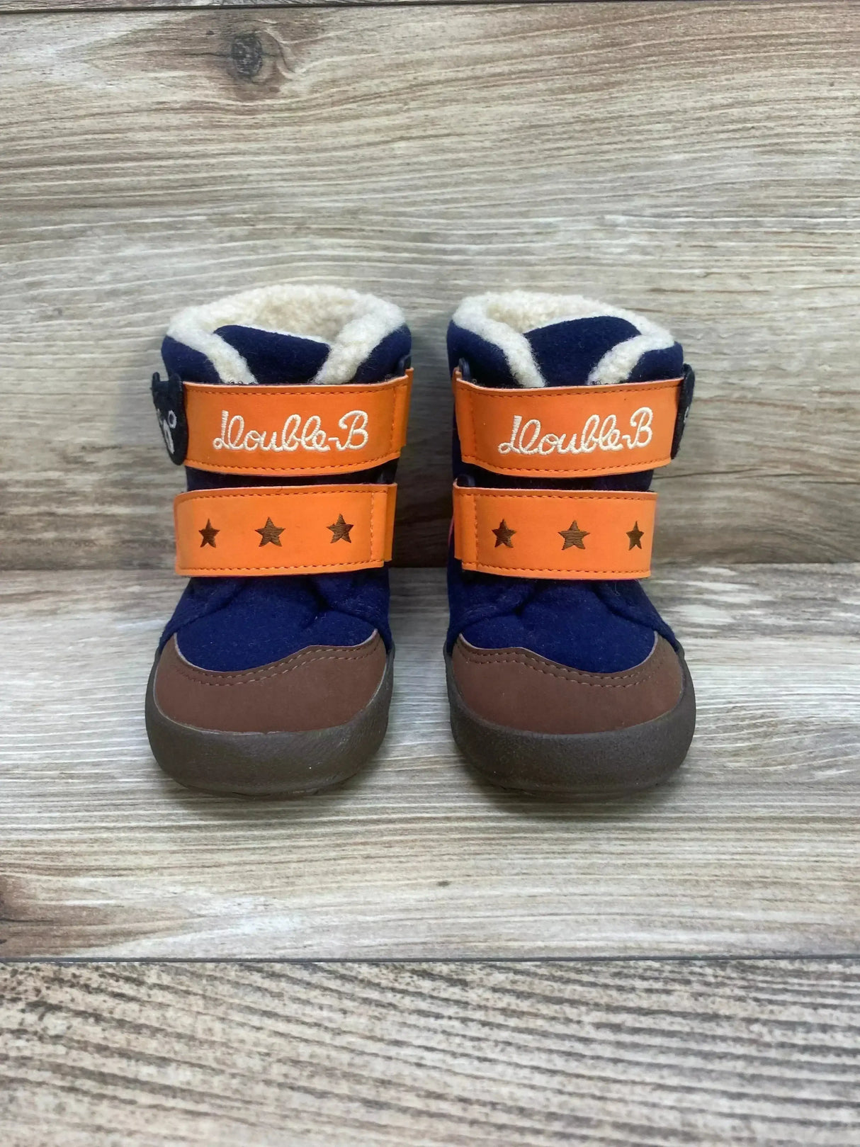 Front view of Double B. miki House toddler winter boots with orange star straps and faux shearling lining on wood background