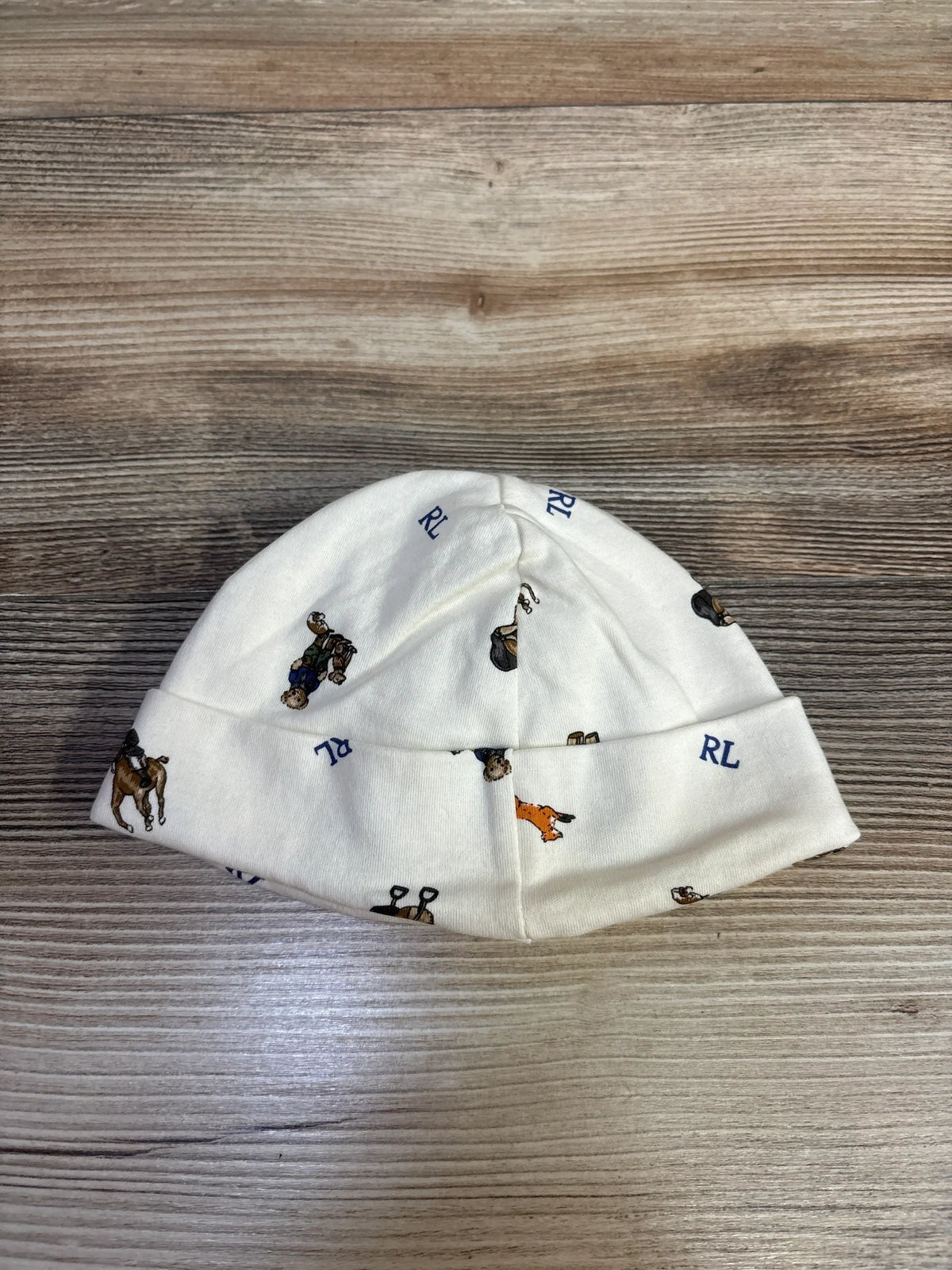 Front view of Ralph Lauren ivory fold-over cap for ages 0 to 9 months, showcasing printed motifs and cuff on wood
