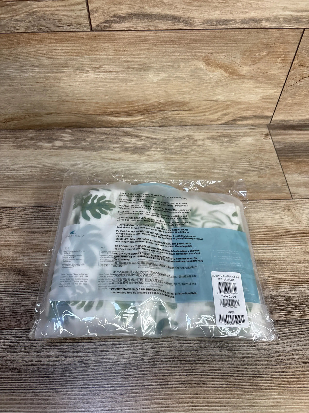 Top-down of folded Little Unicorn muslin sleep sack (6-12M) in clear packaging showing green tropical leaf print on wood