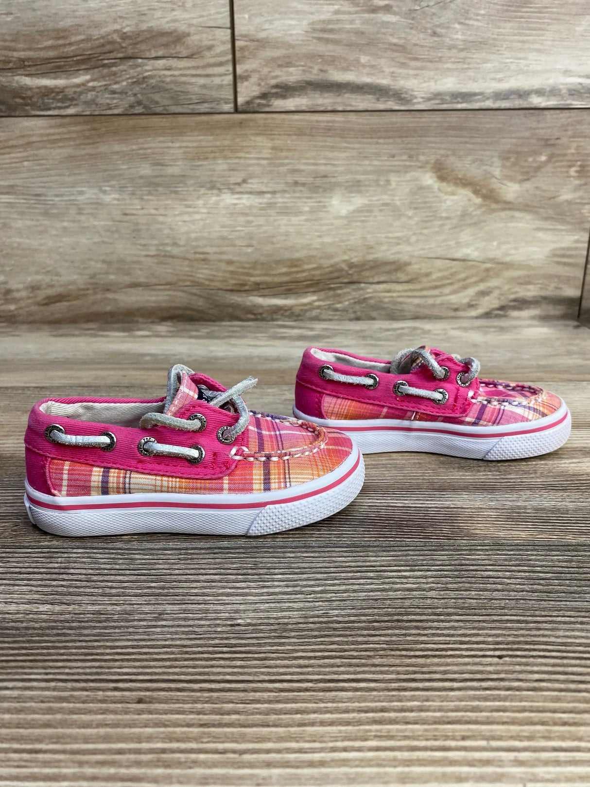 Side view of Sperry Top-Sider pink plaid boat shoes showing moc-toe stitching, silver eyelets and white rubber sole on wood background