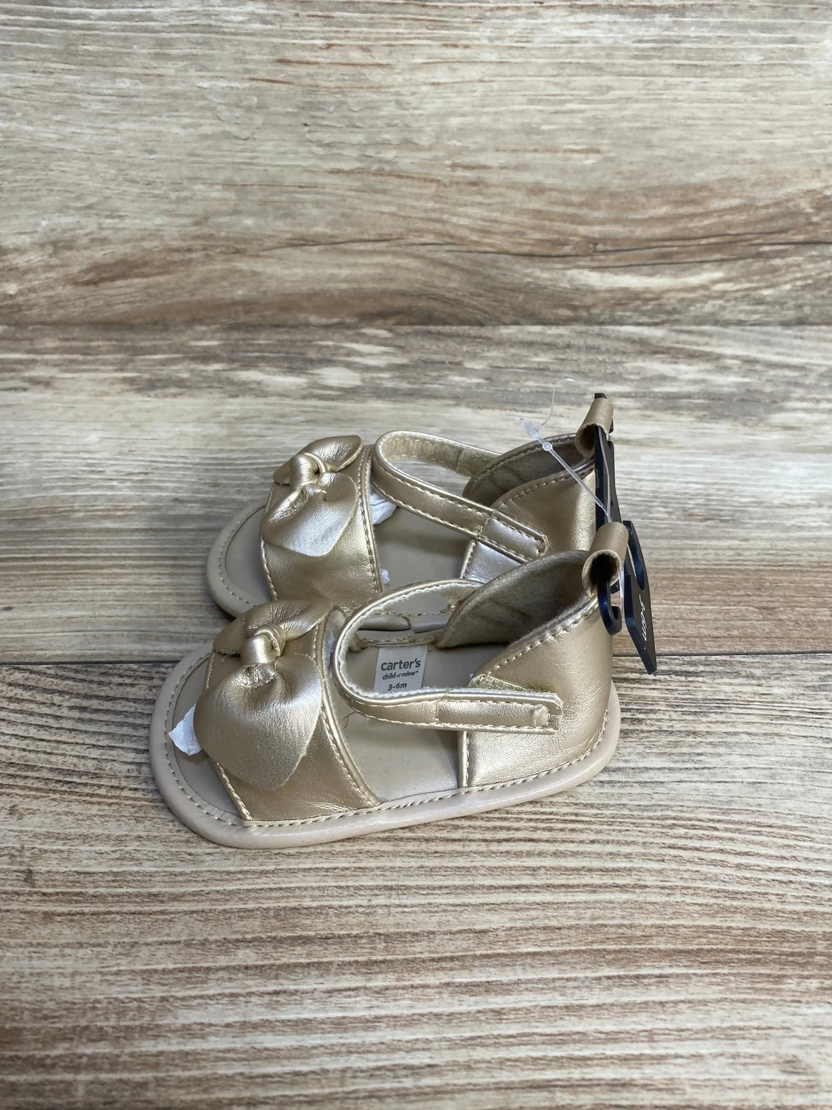 Angled top view of a pair of gold baby sandals showcasing bow detail and velcro ankle strap, size 3-6 months, on wood background