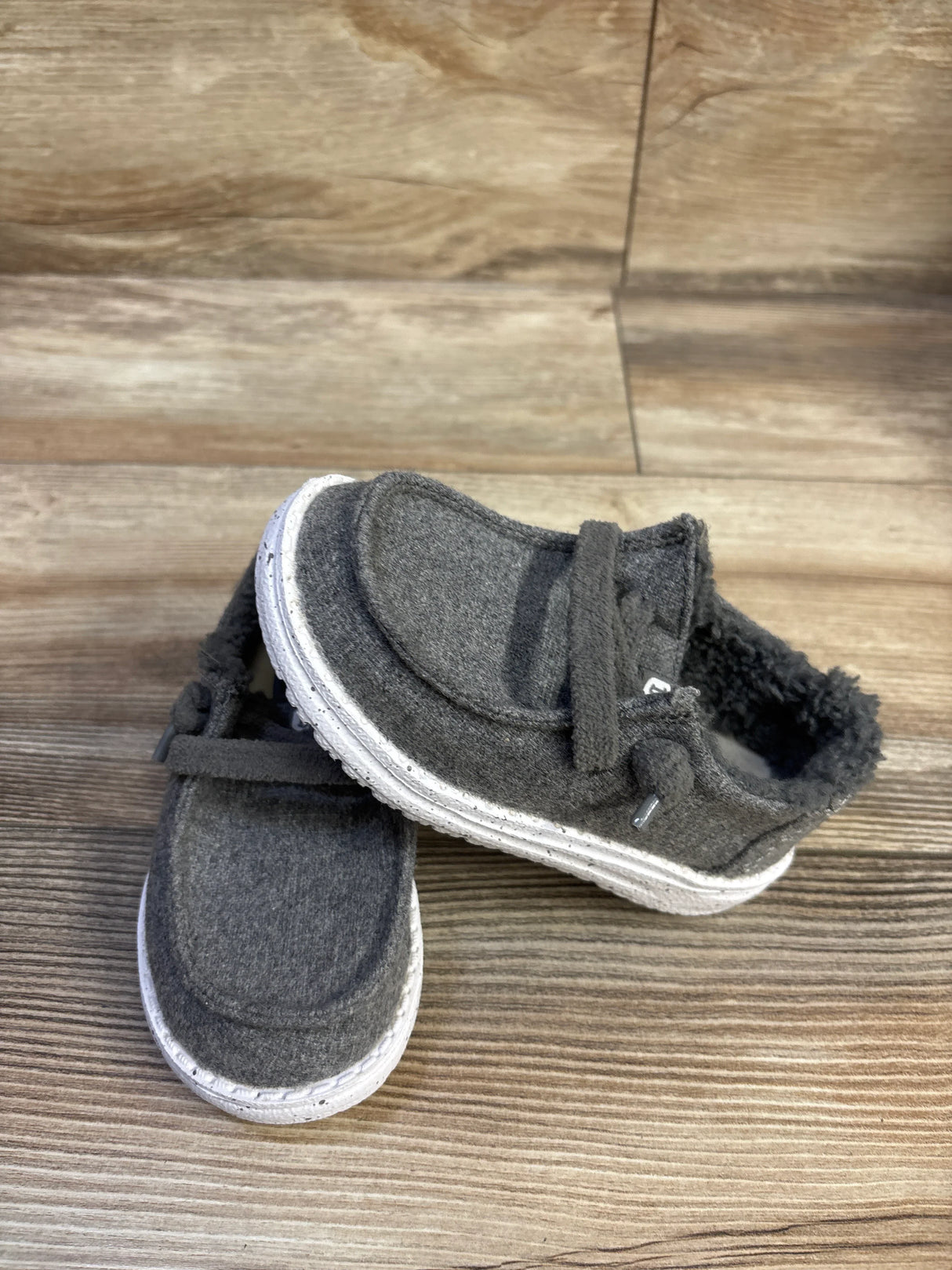 Angled view of Hey Dude Wendy kids grey wool slip-on shoe showcasing sherpa lining and white speckled sole on wood background