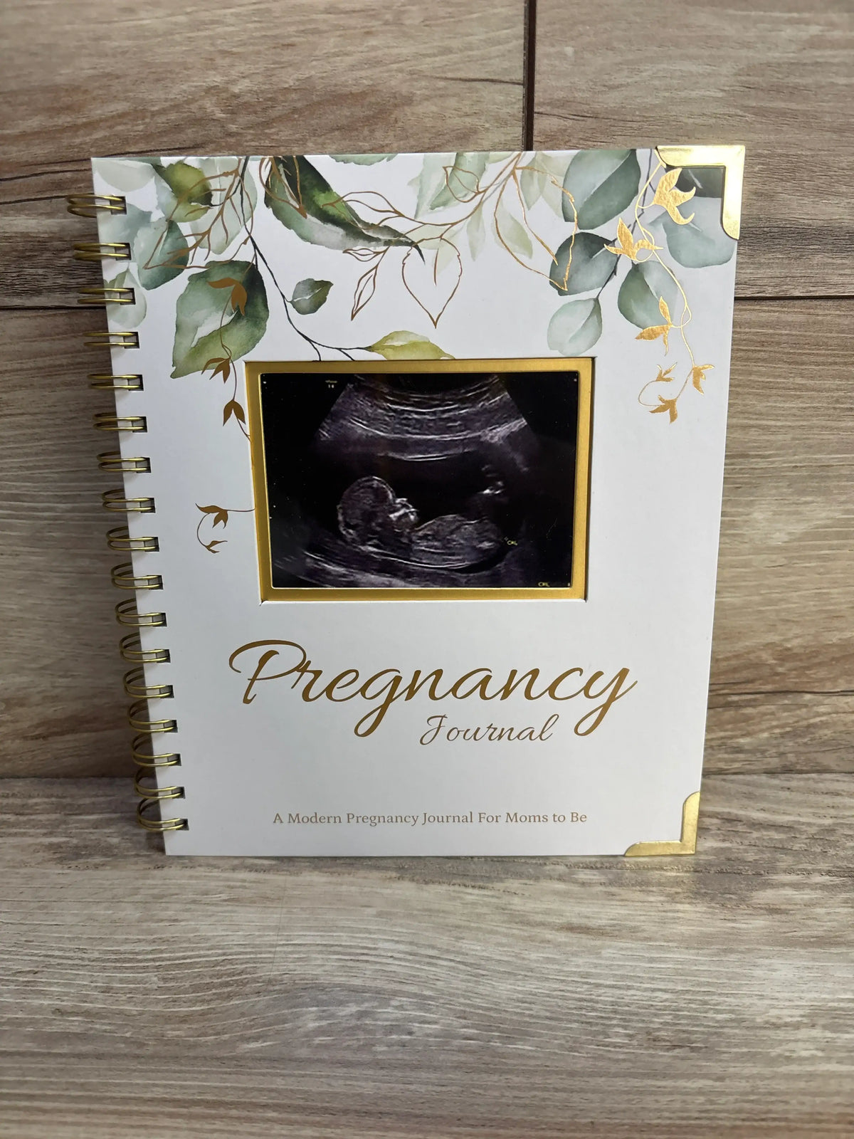 Front view of KeaBabies Inspire Pregnancy Journal showcasing ring binding, foliage cover, and ultrasound photo on a wood surface