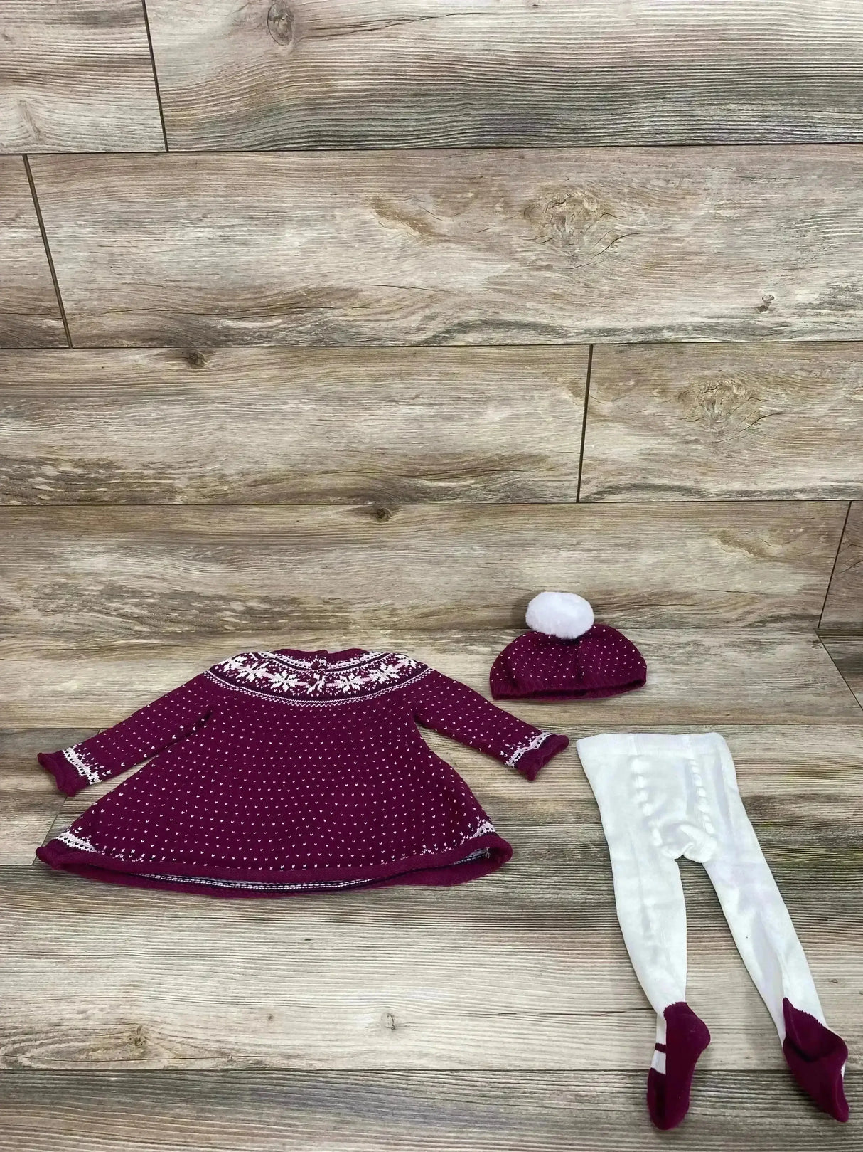 Flat lay of three-piece Fair Isle knit dress set showcasing purple dress with white patterned yoke, matching pom hat, and white footed tights on wood background