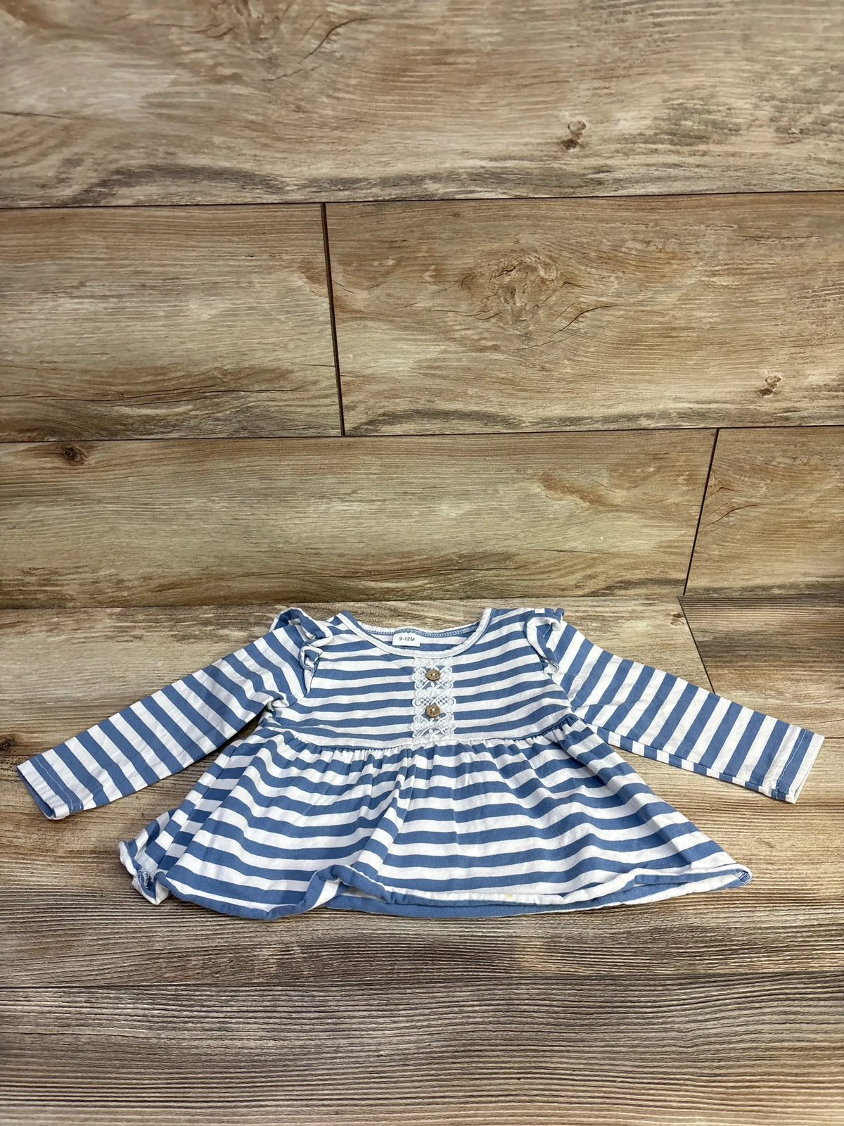 Front view of Durtebeua blue striped peplum henley top with long sleeves, button placket, and peplum hem on wood background