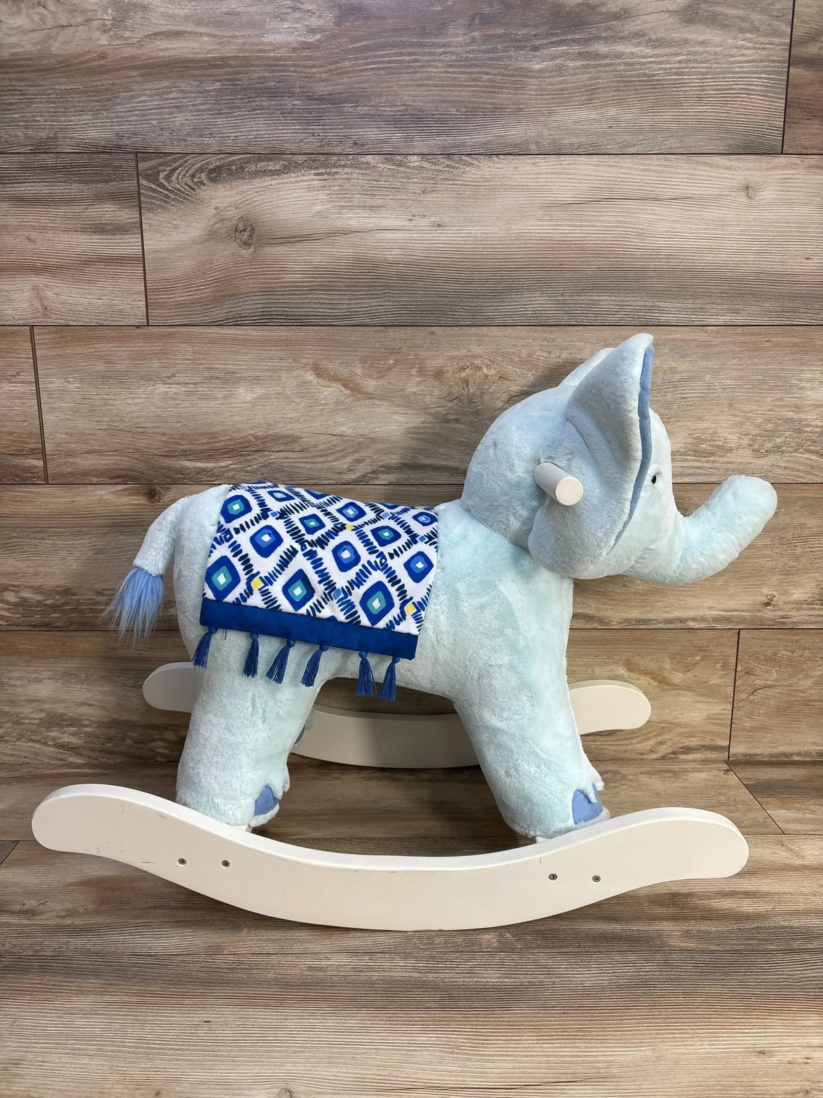 Side view of elephant nursery rocker showing pale blue plush body, blue patterned saddle with tassels on wood background
