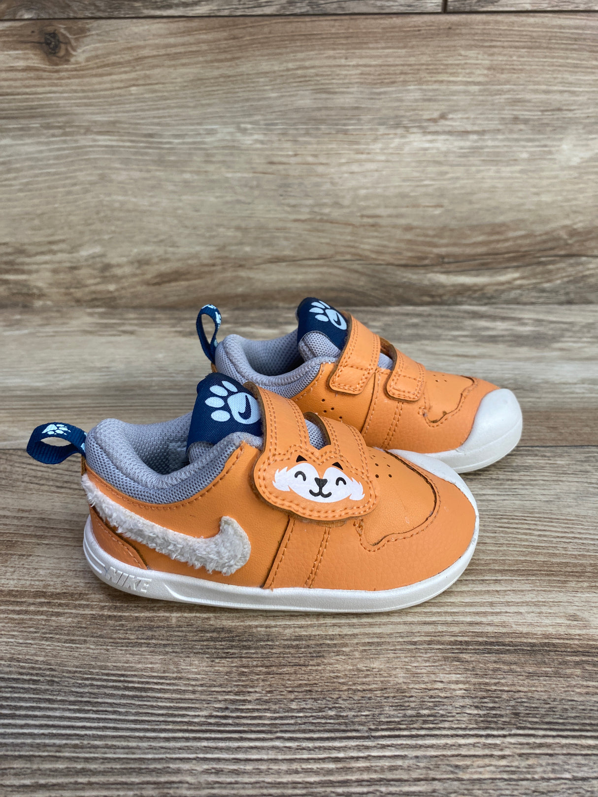 Orange toddler Nike shoes with white fuzzy swoosh and smiling animal face on velcro strap on wood floor