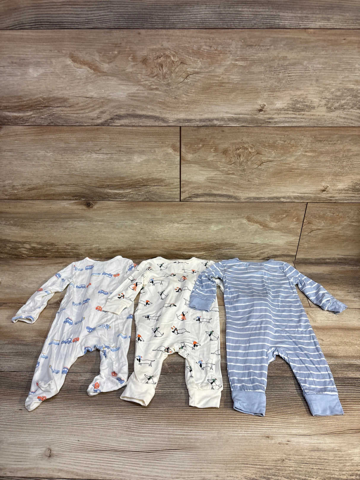 Flat lay of three Little Me bamboo footless sleepers size 3M, ivory and prints cars, dogs and blue stripes on wood backdrop