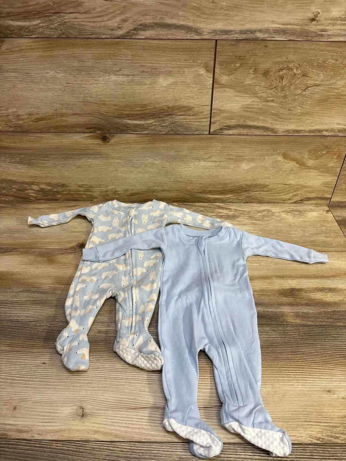 Top view of two newborn footed sleepers, front zips and non slip soles, solid blue and bear print light blue on wood floor