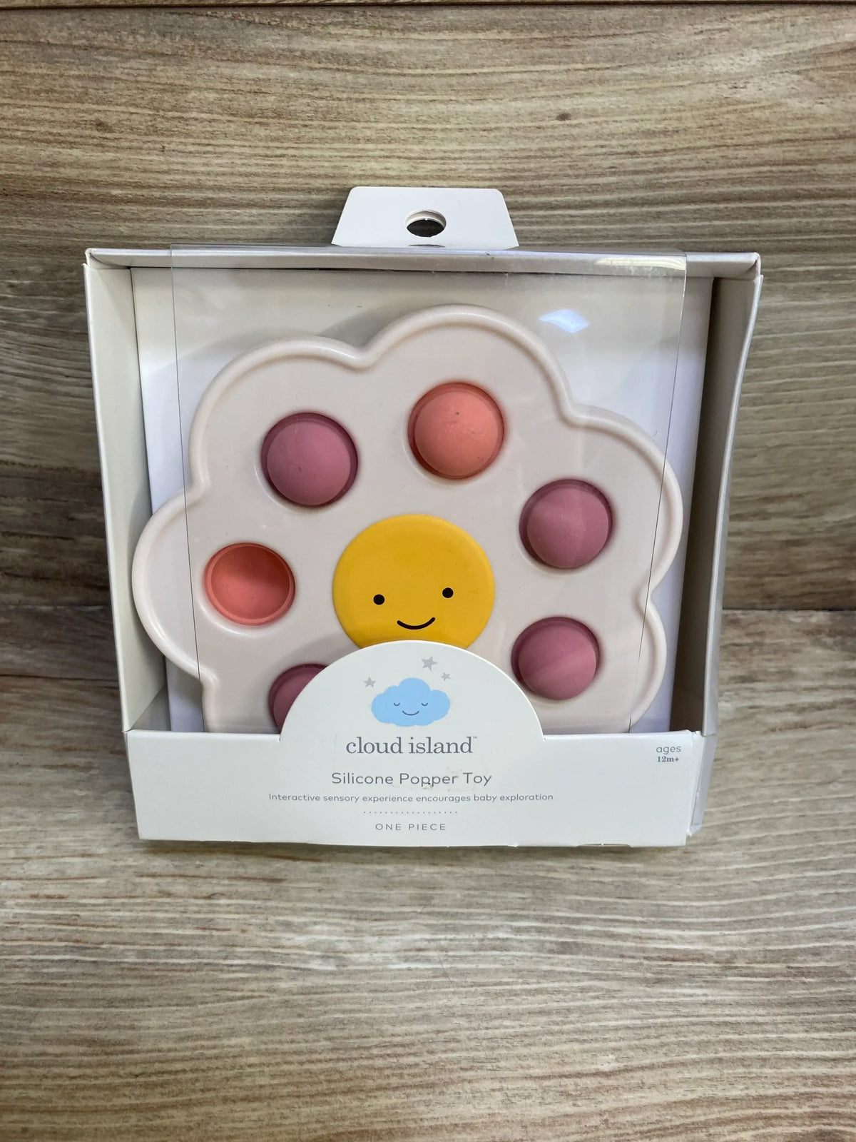 Front view of Cloud Island silicone popper toy in box, flower-shaped with seven pink and coral poppable bubbles and a smiling yellow center on wood background