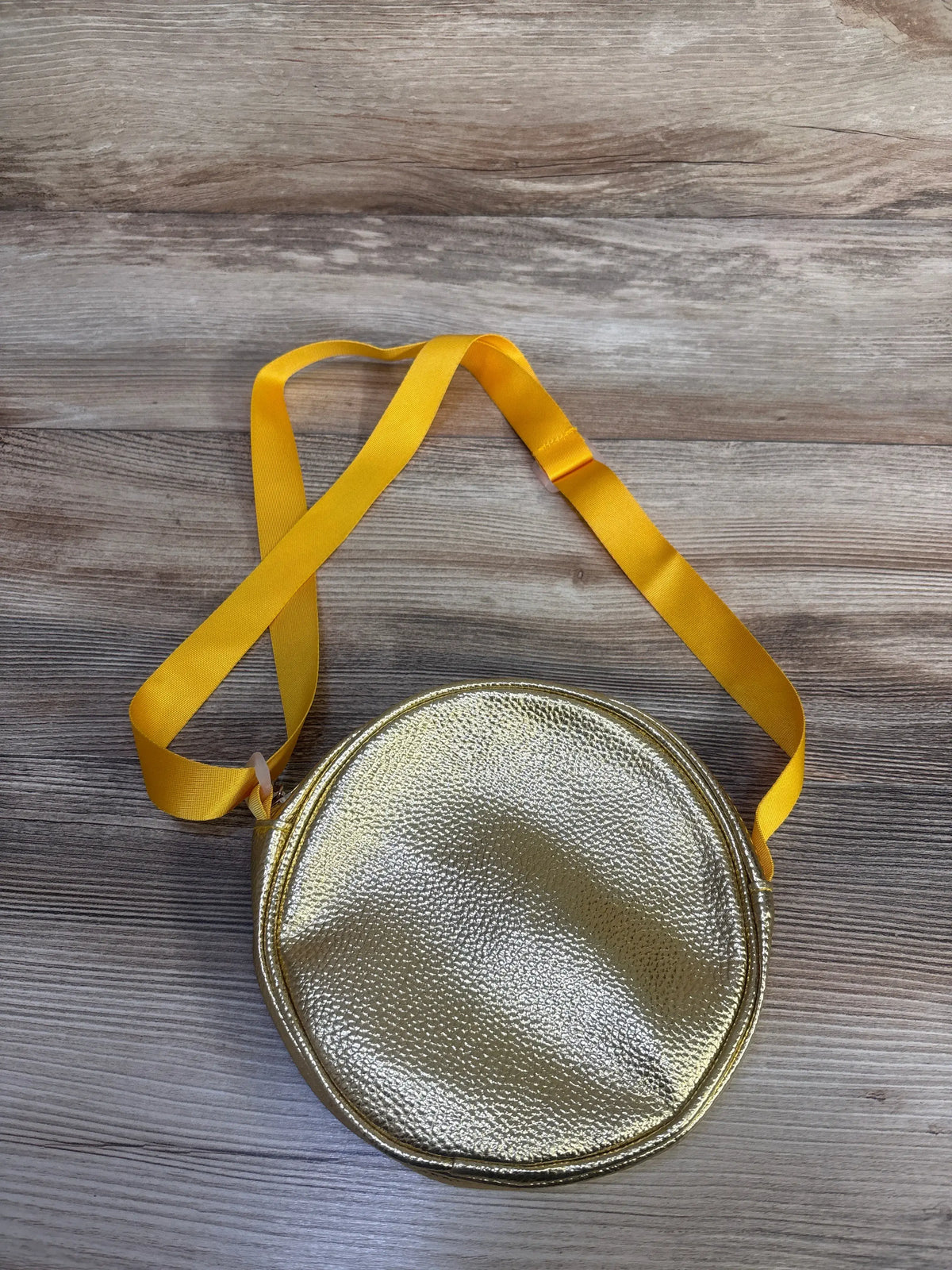 Top view of a gold round purse showcasing a textured metallic finish and long yellow shoulder strap on a wood-grain surface